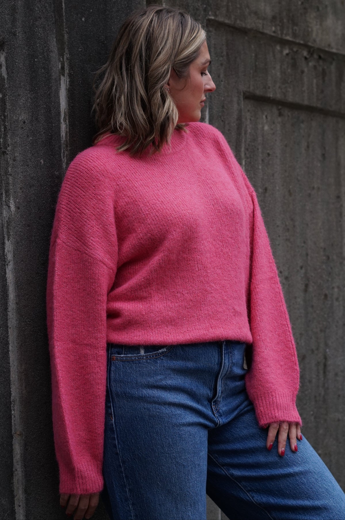 A woman wearing a fuzzy pink knit sweater and blue jeans leans against a textured concrete wall.