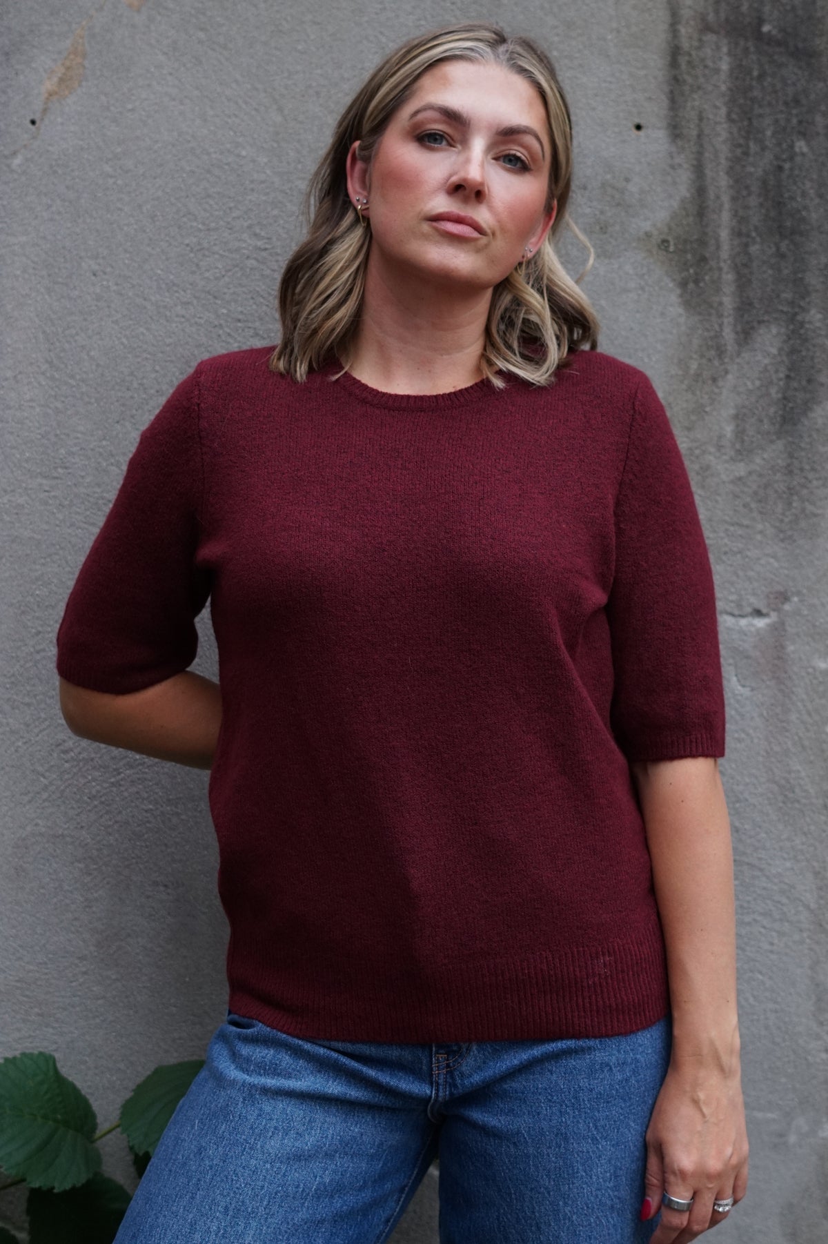 A woman wearing a short-sleeve crewneck knit sweater in deep burgundy, paired with blue jeans, standing against a textured gray concrete wall.
