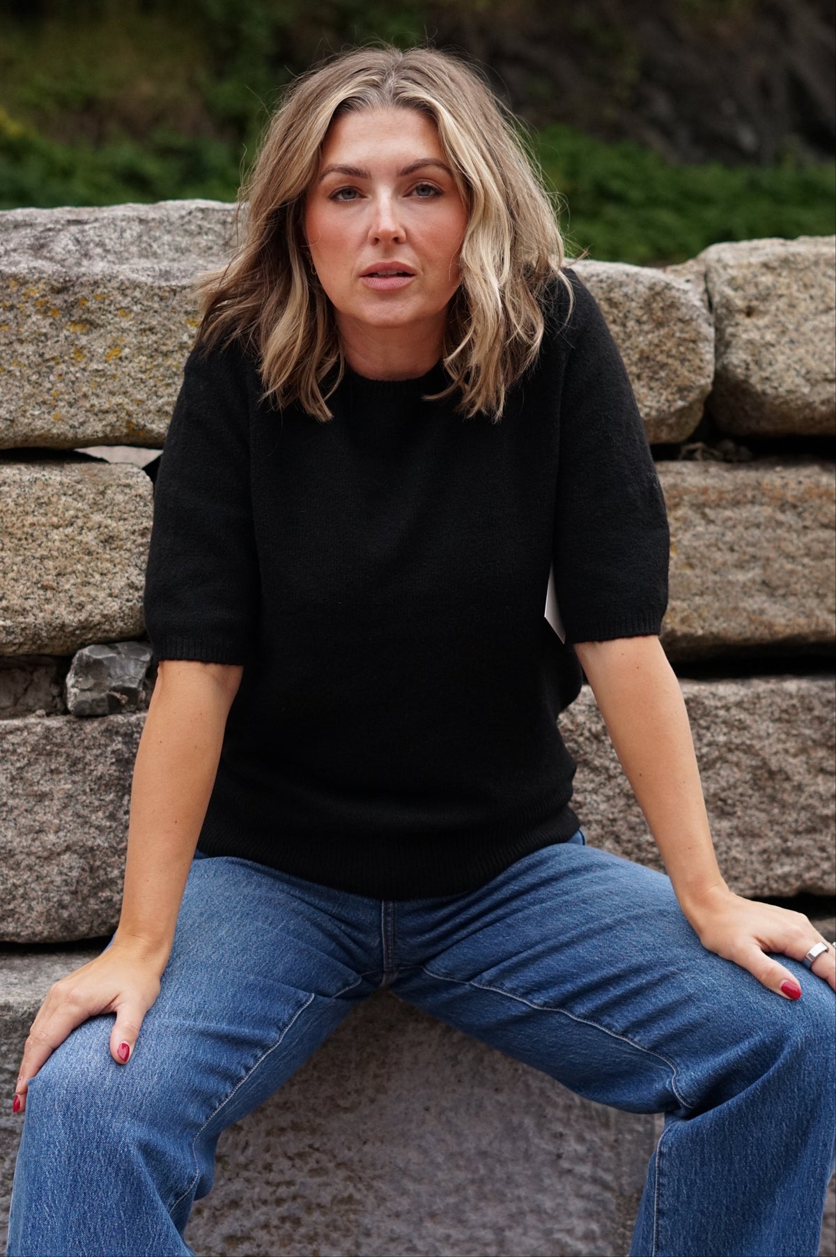 A woman with blonde hair sitting on a stone wall, wearing a black short-sleeve top and blue jeans.