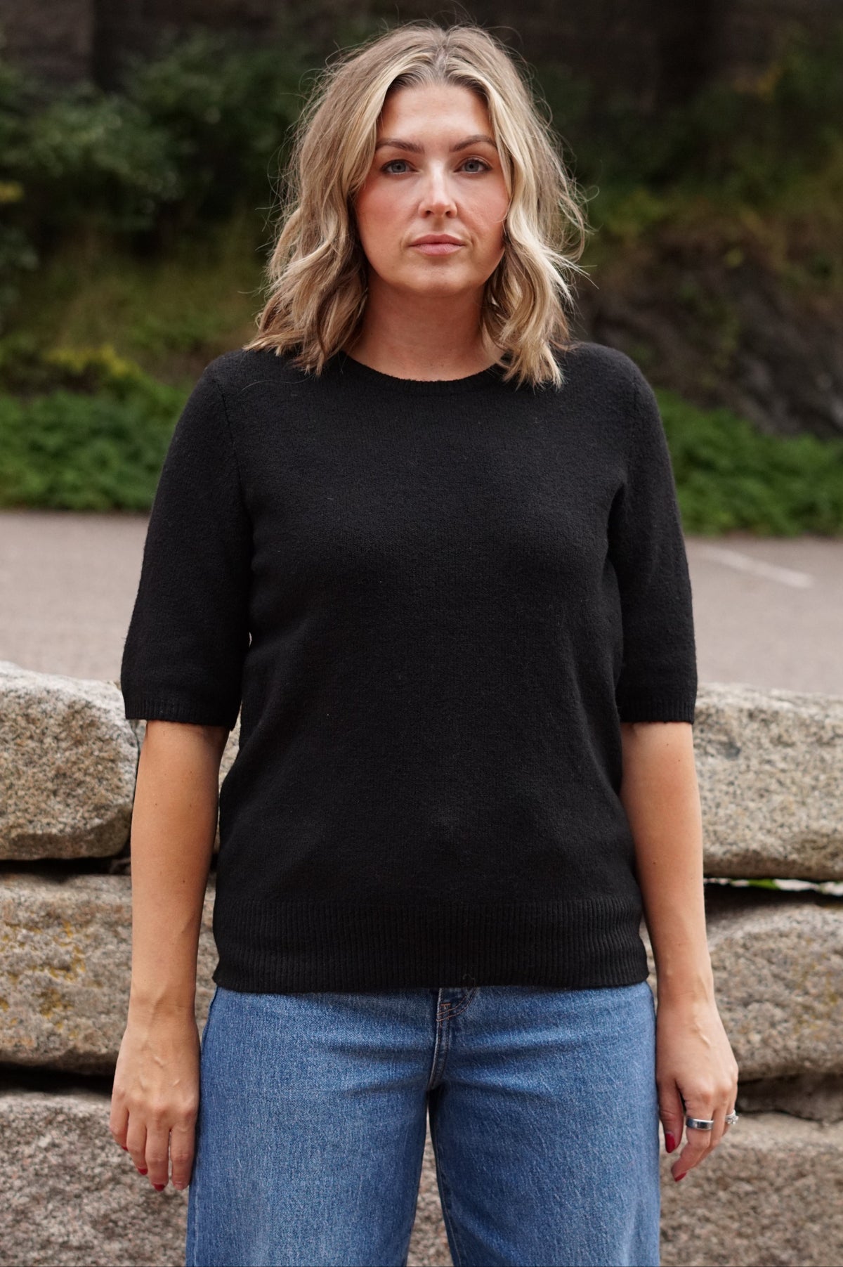 A woman with blonde hair wearing a black short-sleeve crewneck knit sweater and blue jeans, standing outdoors in front of a stone wall and greenery.