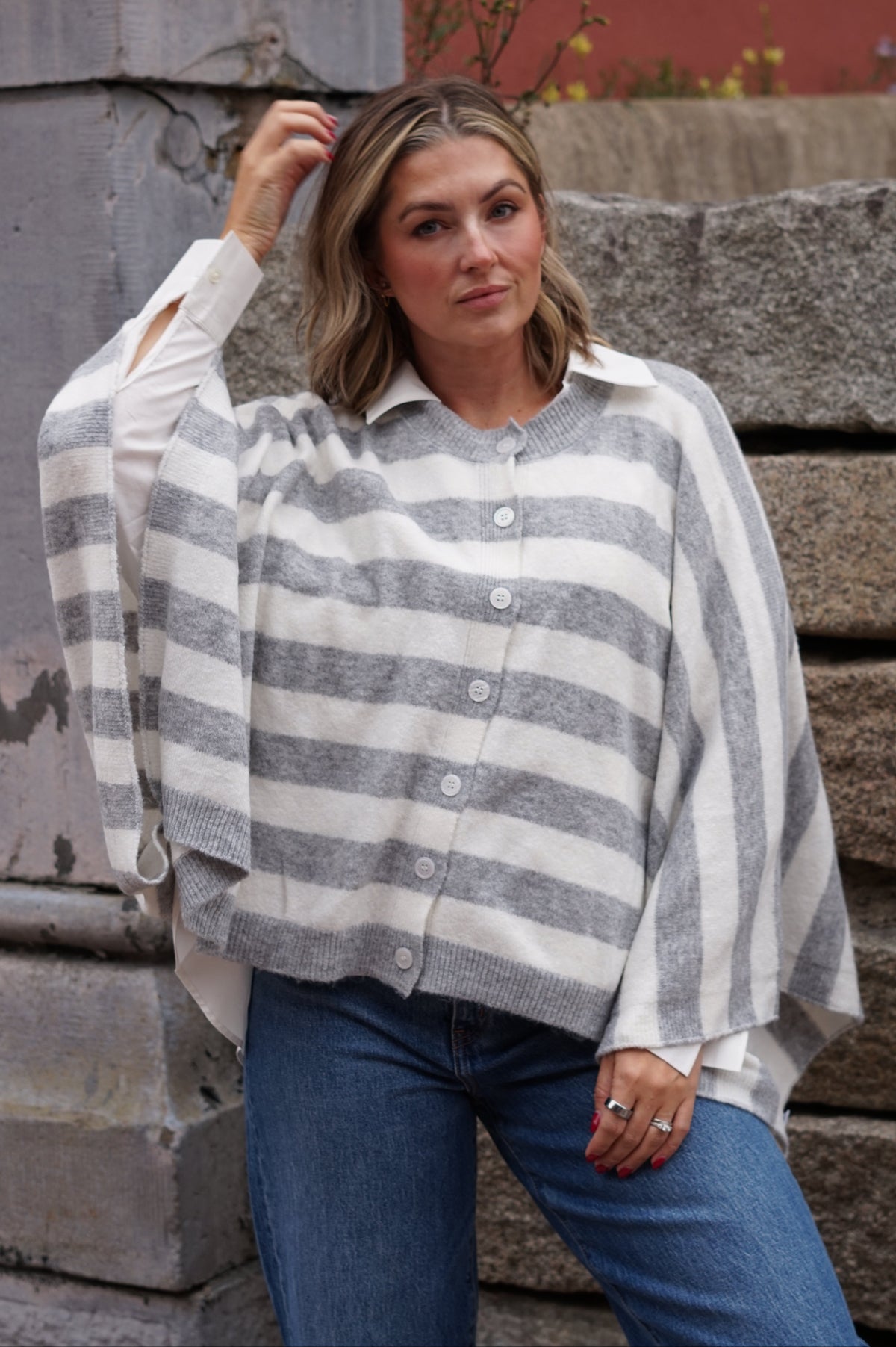 A woman wearing a gray and white striped button-up cardigan with wide sleeves, paired with blue jeans, posing outdoors against a stone wall.