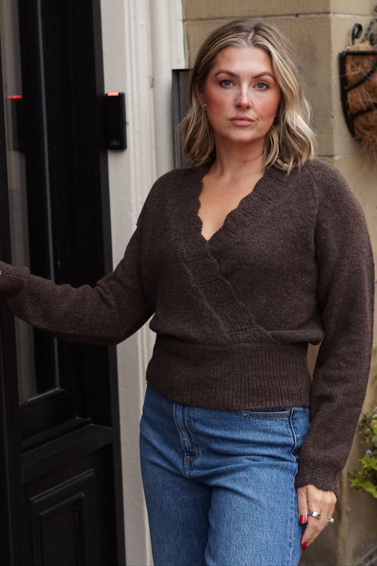 A woman wearing a brown knit V-neck sweater and blue jeans, standing in front of a dark door and stone wall.