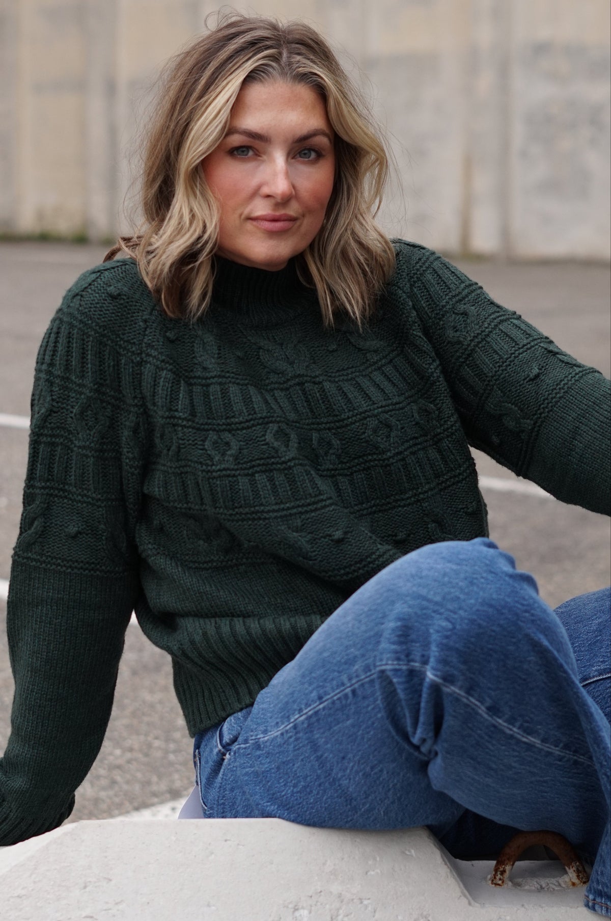 A woman wearing a dark green cable-knit turtleneck sweater and blue jeans sits on a concrete ledge in an outdoor urban setting.
