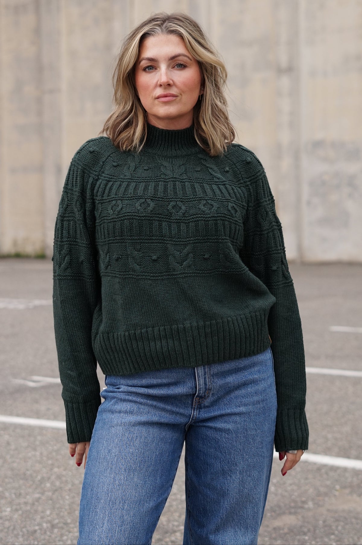 A woman wearing a dark forest green cable-knit mock neck sweater and blue jeans, standing in an outdoor paved area with a concrete wall in the background.
