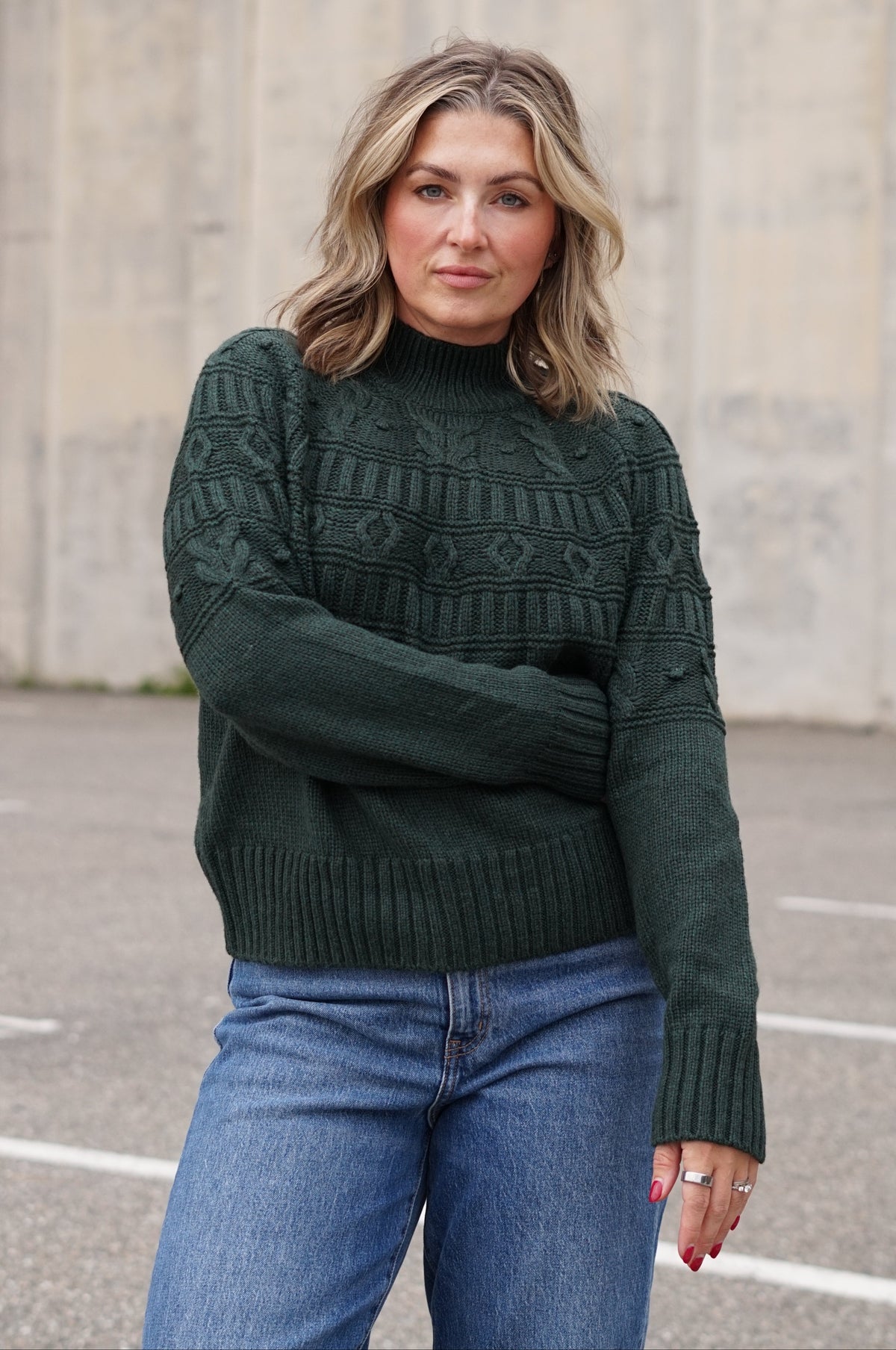 A woman wearing a dark forest green cable-knit mock-neck sweater and blue jeans, standing outdoors against a concrete wall.