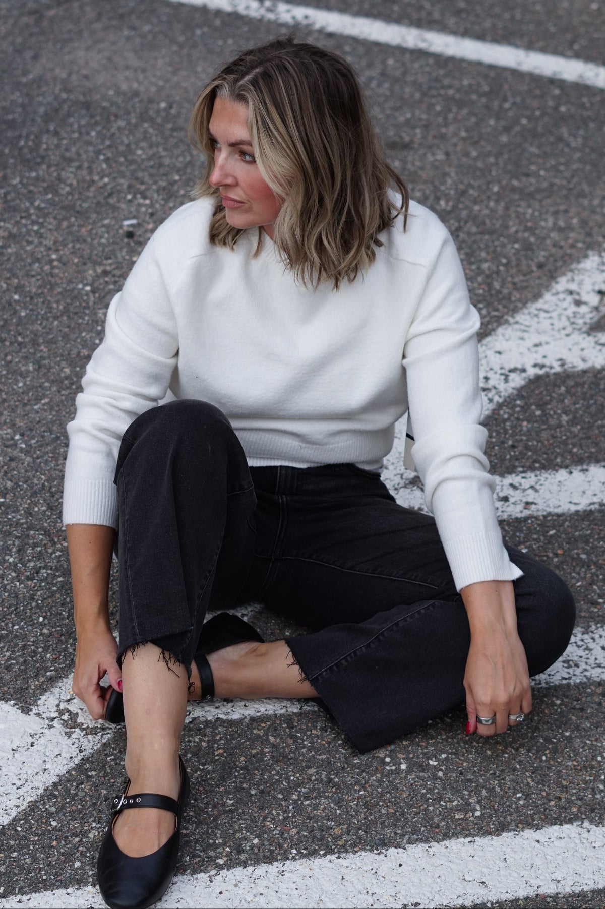 A woman sits on asphalt at a crosswalk wearing a white knit mock-neck sweater, black cropped jeans, and black Mary Jane shoes.