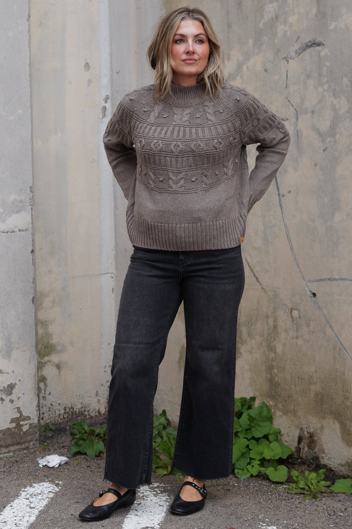 A woman stands outdoors against a weathered concrete wall wearing a taupe knit mock-neck sweater with a geometric pattern, black wide-leg jeans, and black flats.