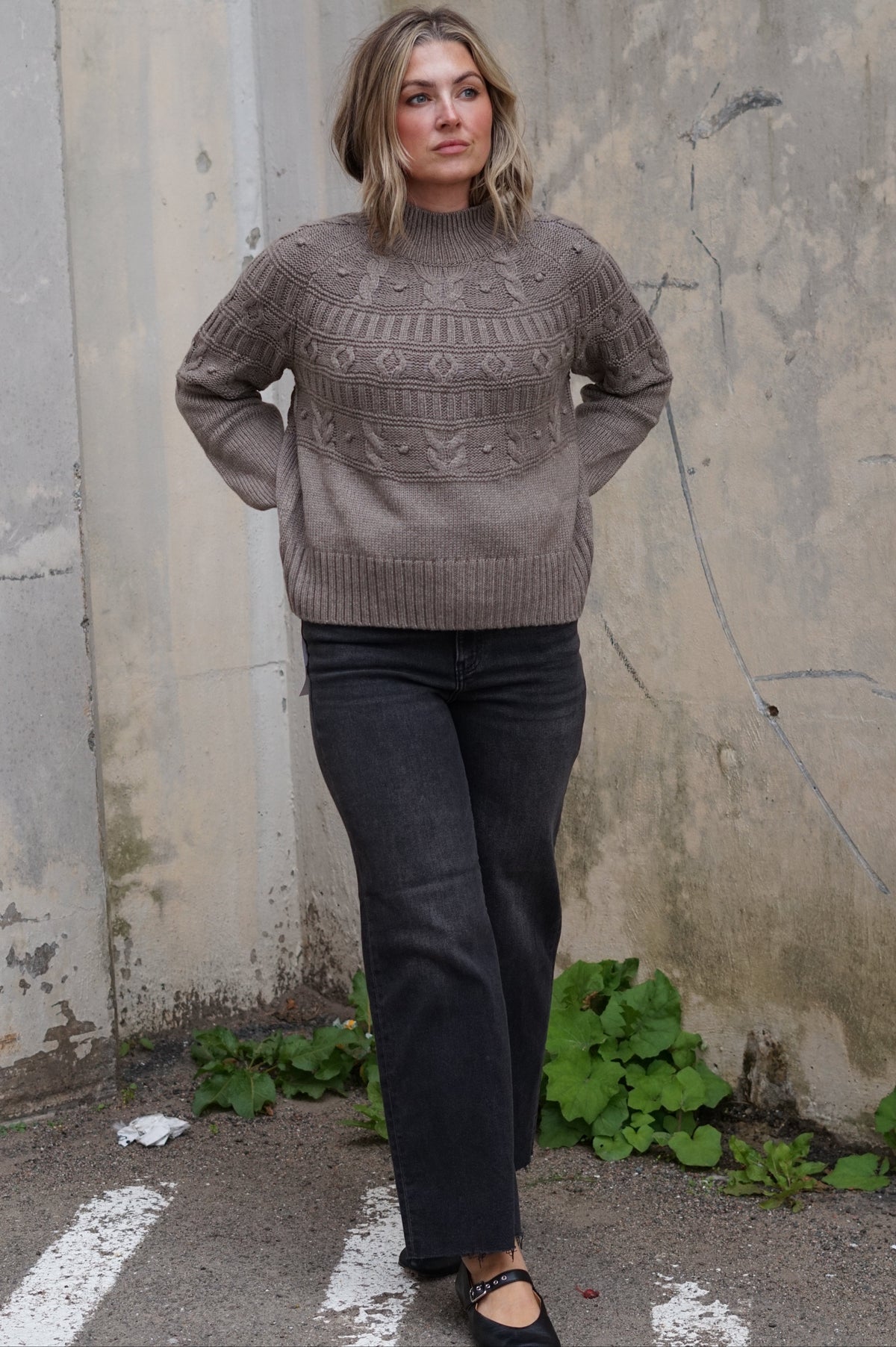 A woman wearing a taupe-brown cable-knit mock-neck sweater, dark charcoal wide-leg jeans, and black Mary Jane flats, standing against a weathered concrete wall.