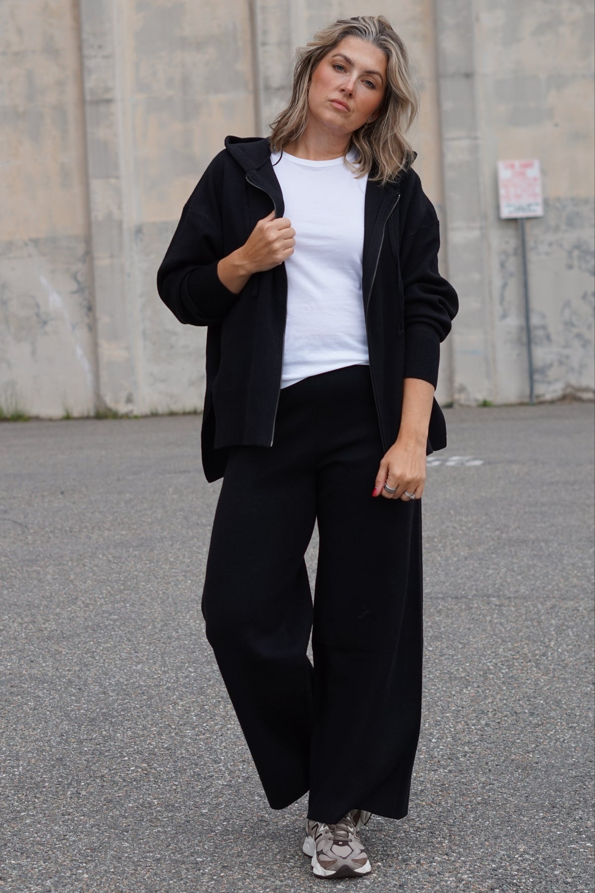 A woman wearing a black zip-up hoodie, white t-shirt, and black wide-leg pants standing outdoors on asphalt in front of a concrete wall.
