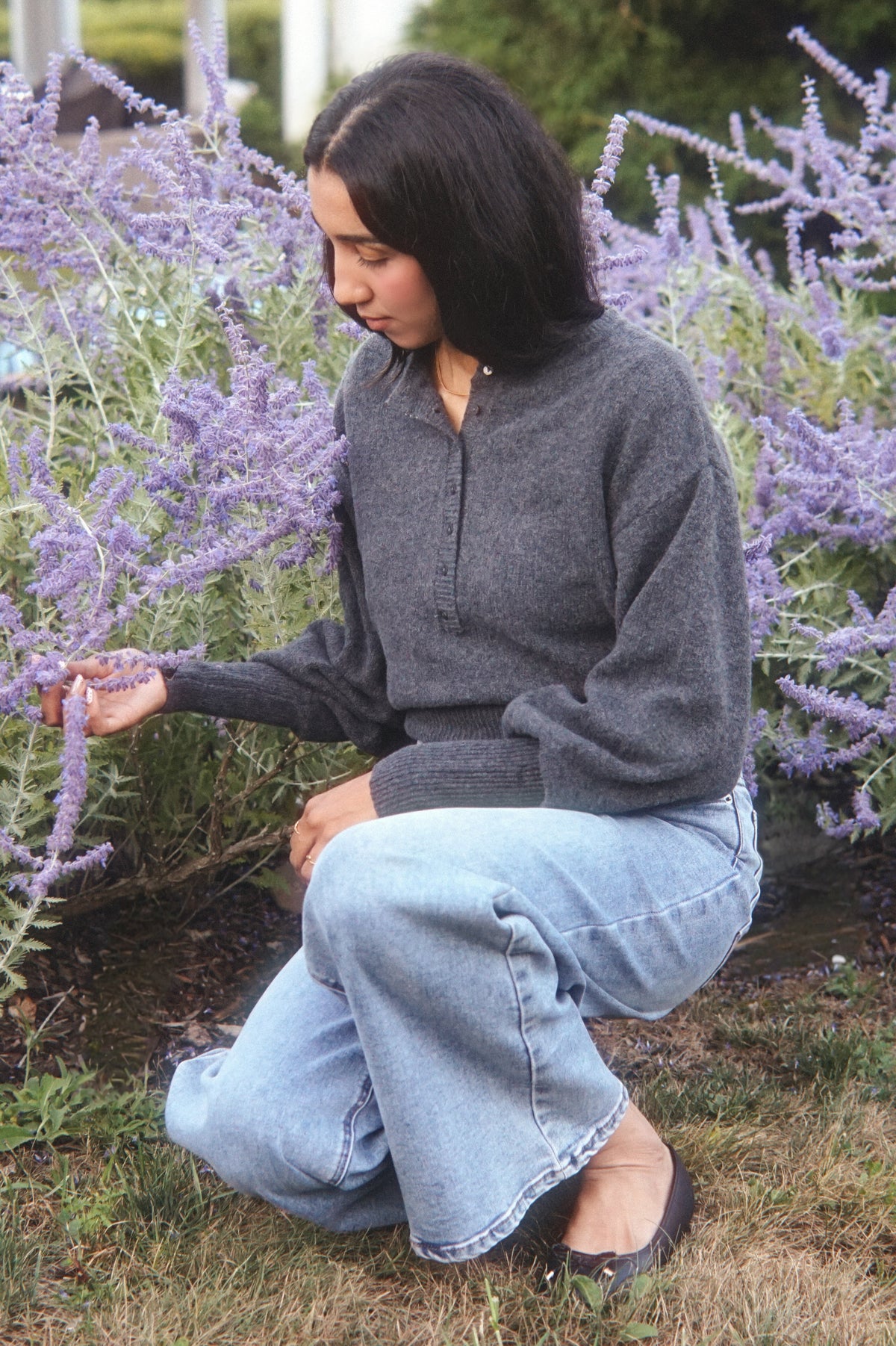 A woman crouching in a garden among tall purple flowers, wearing a dark gray quarter-zip pullover sweater and light blue wide-leg jeans.