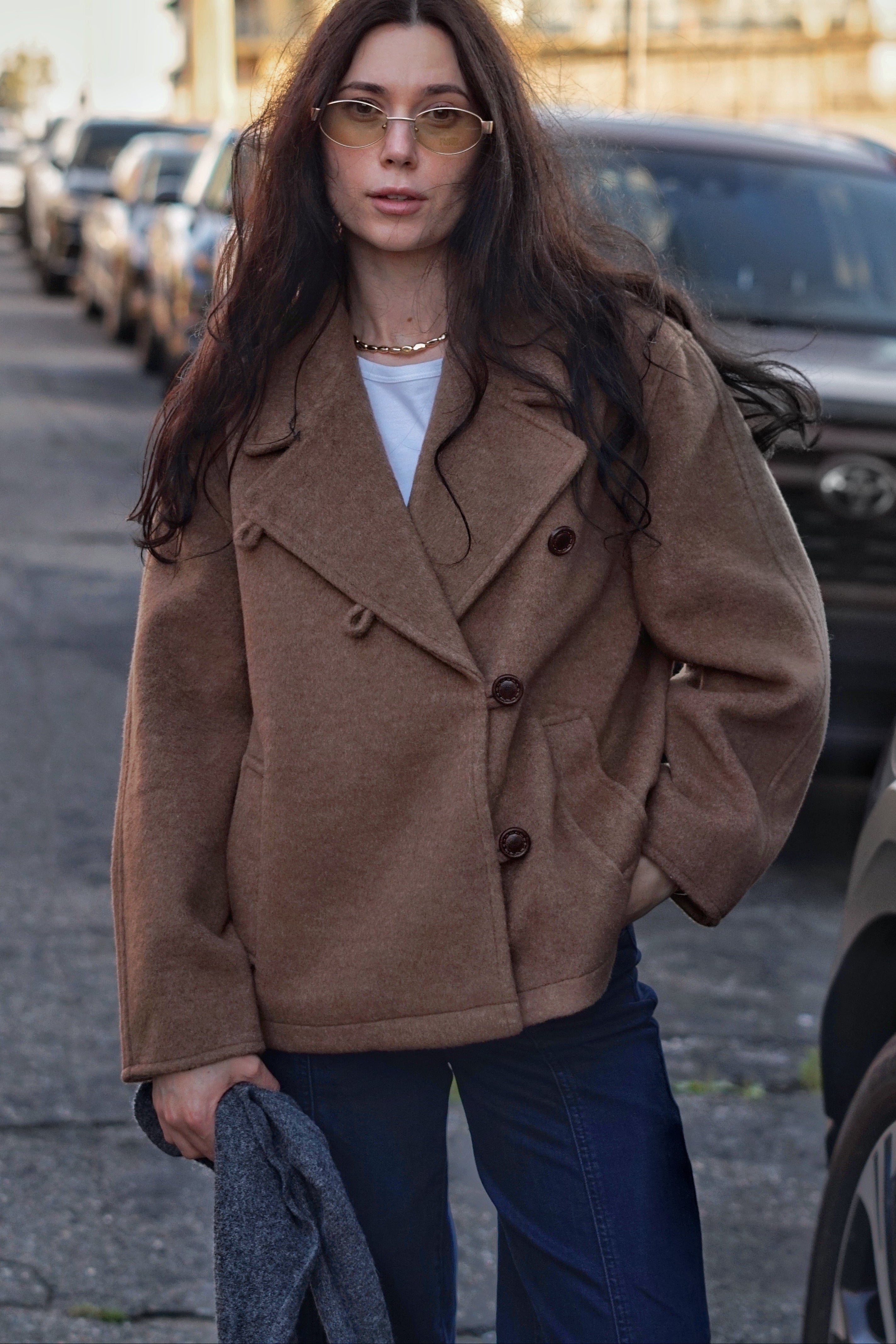 A woman wearing a brown wool-blend double-breasted coat over a white shirt and blue jeans, standing on a street with parked cars in the background.