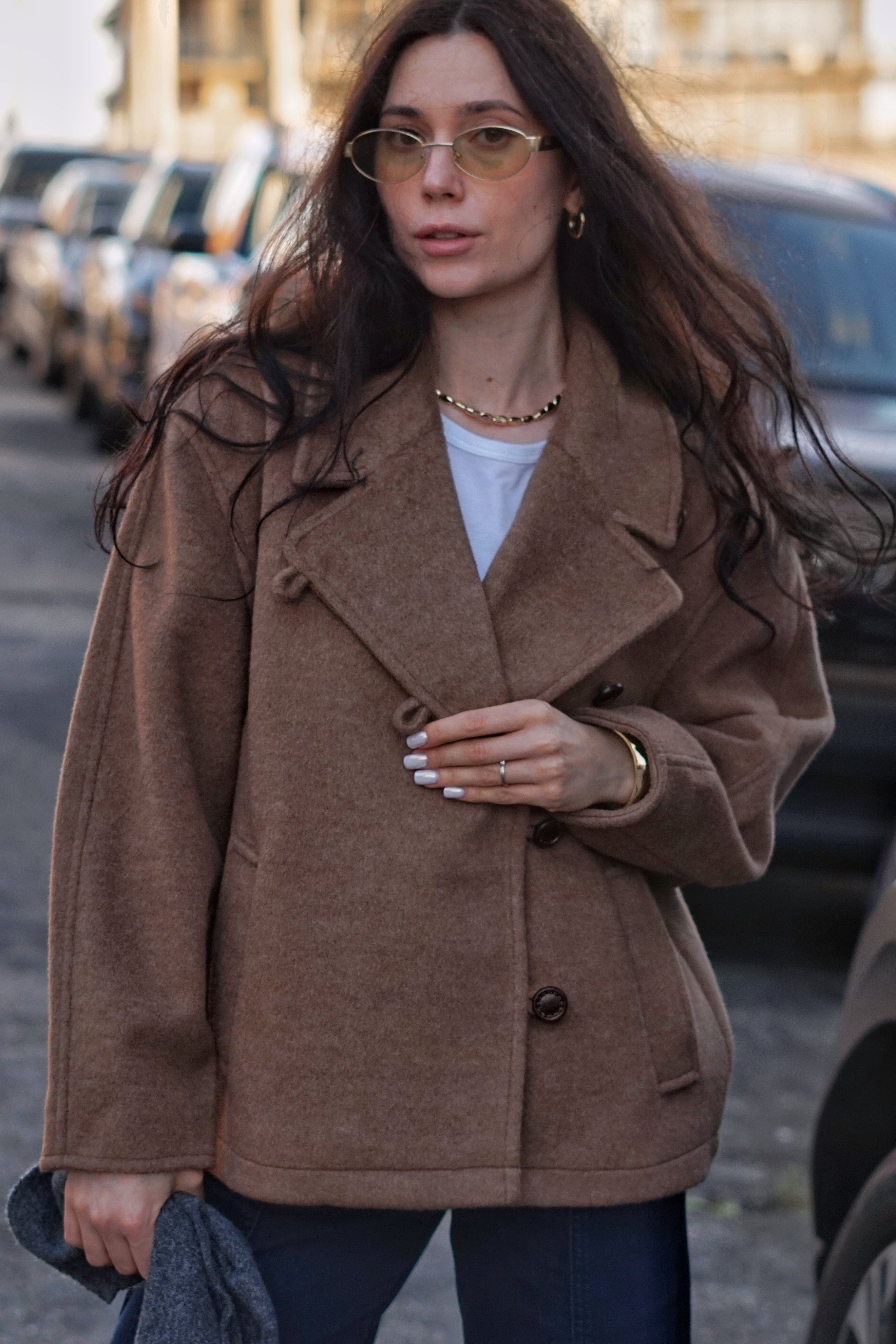 A woman wearing a brown double-breasted coat over a white top and dark blue jeans, standing on a city street with parked cars in the background.