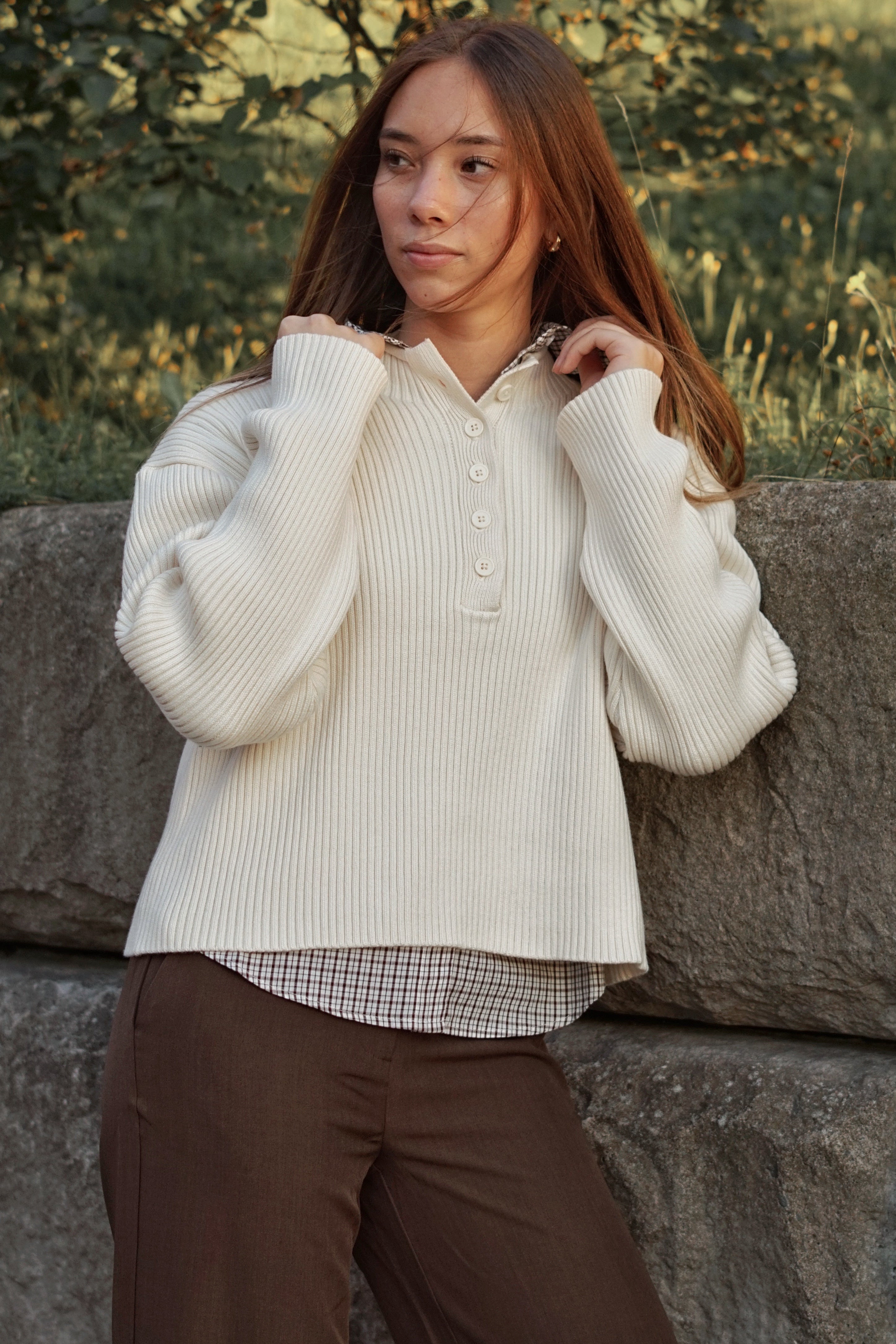 A woman with long light brown hair stands outdoors against a stone wall, wearing a cream ribbed knit button-front sweater over a small checkered shirt and brown pants.