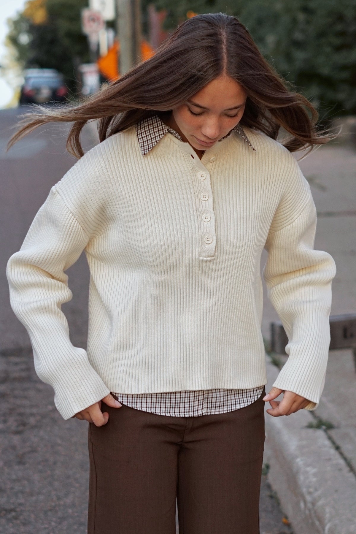 A woman stands on a city street wearing a cream ribbed knit polo-style sweater over a small checkered shirt, paired with brown wide-leg pants.