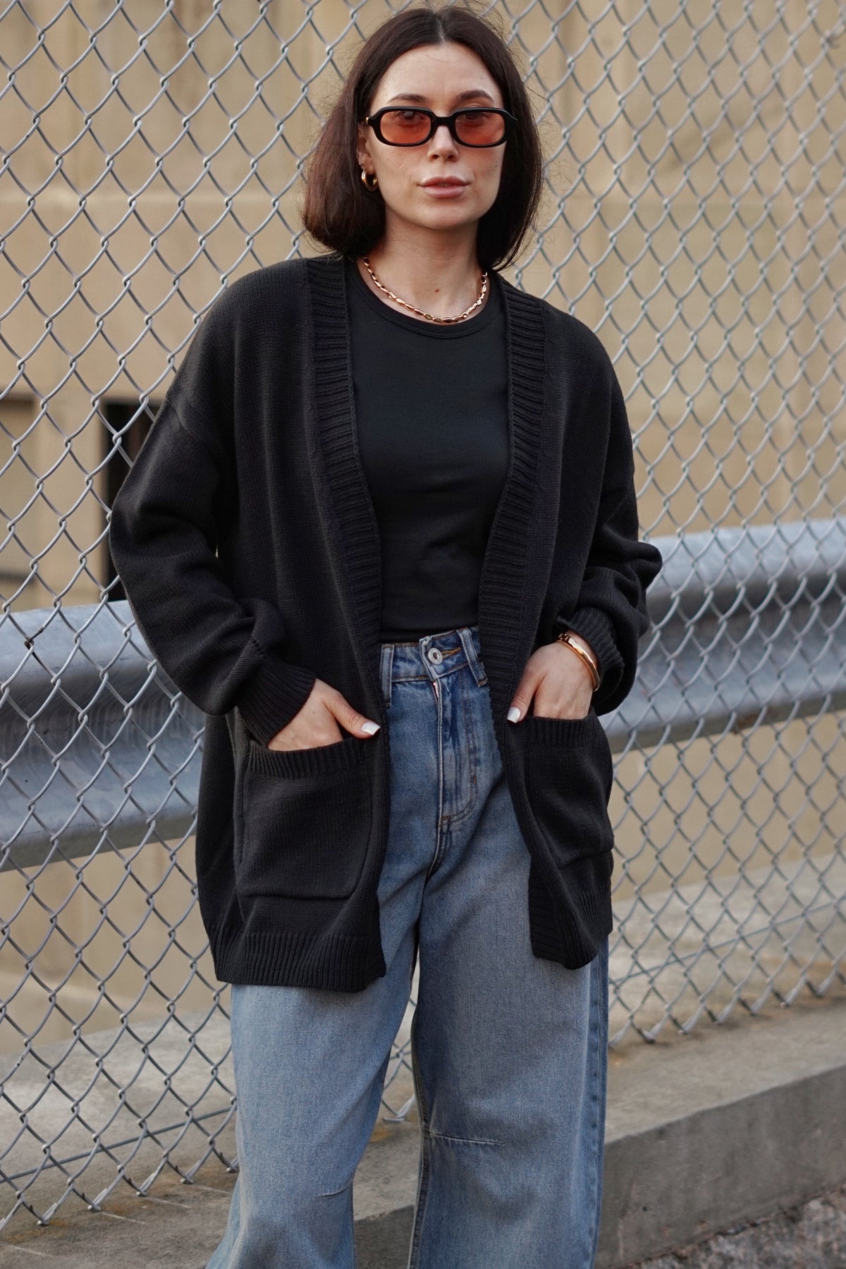 A woman stands outdoors in front of a chain-link fence wearing a black knit cardigan over a black top, light blue wide-leg jeans, and orange-tinted sunglasses.