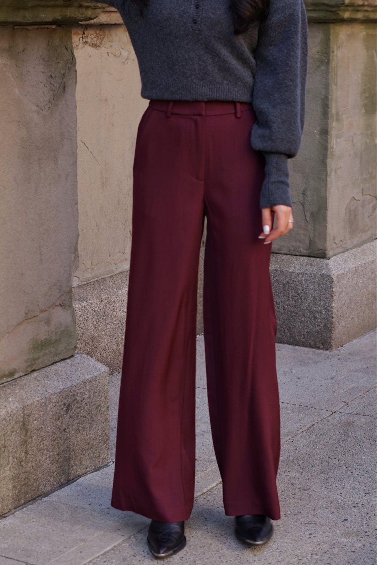 A person wearing burgundy wide-leg trousers with a matching belt, paired with a dark gray knit sweater and black shoes, standing outdoors against a stone wall.