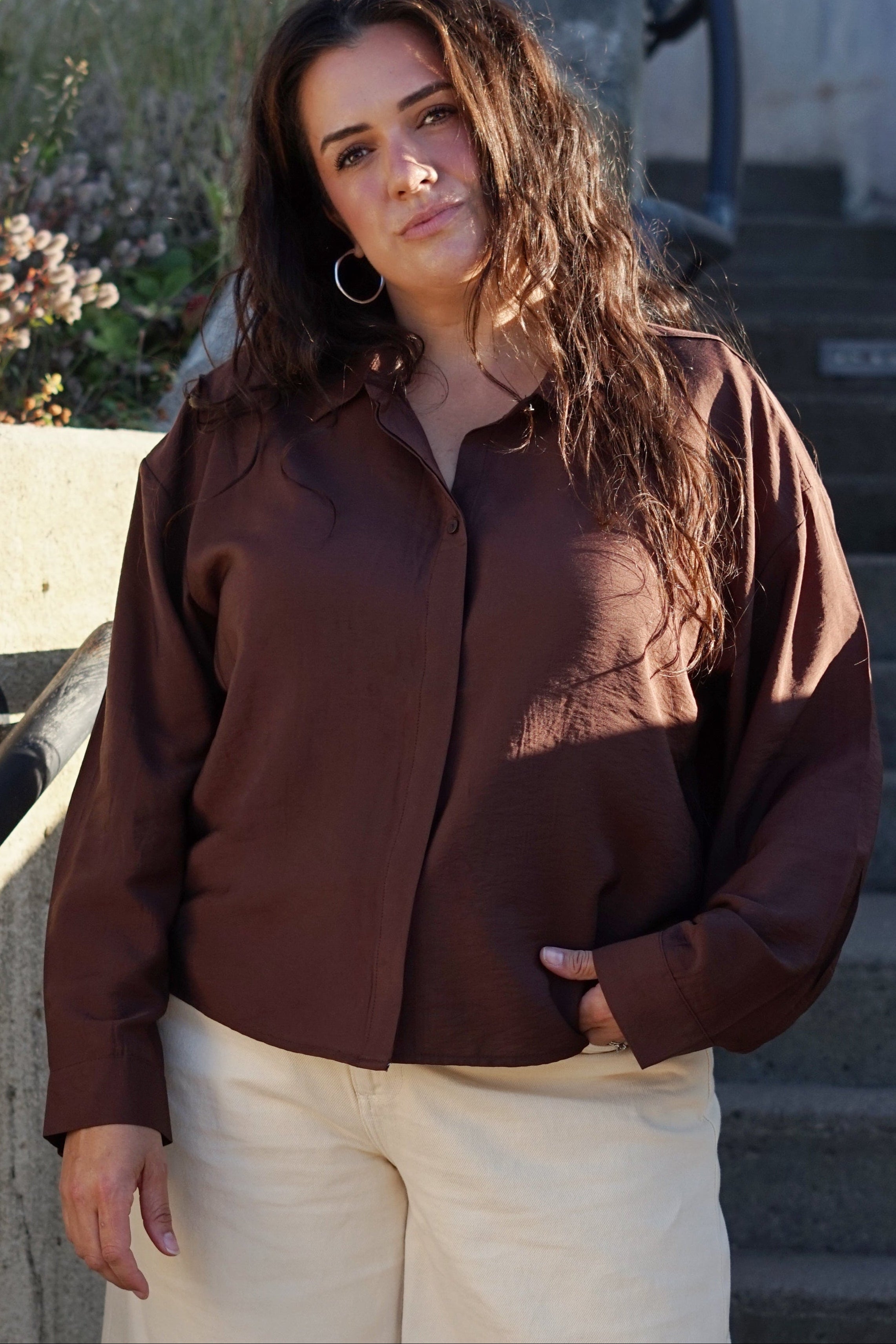 A woman stands on outdoor concrete stairs wearing a dark brown long-sleeve button-up shirt and off-white shorts.