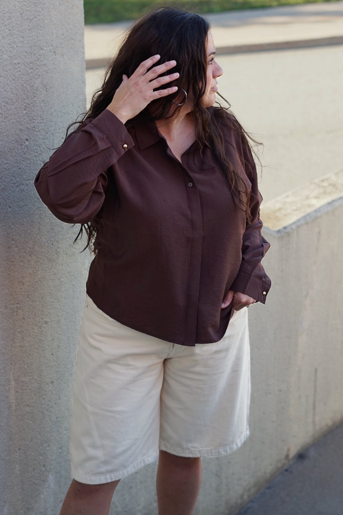 A woman wearing a dark brown long-sleeve button-up shirt and cream-colored shorts, standing outdoors next to a concrete pillar.