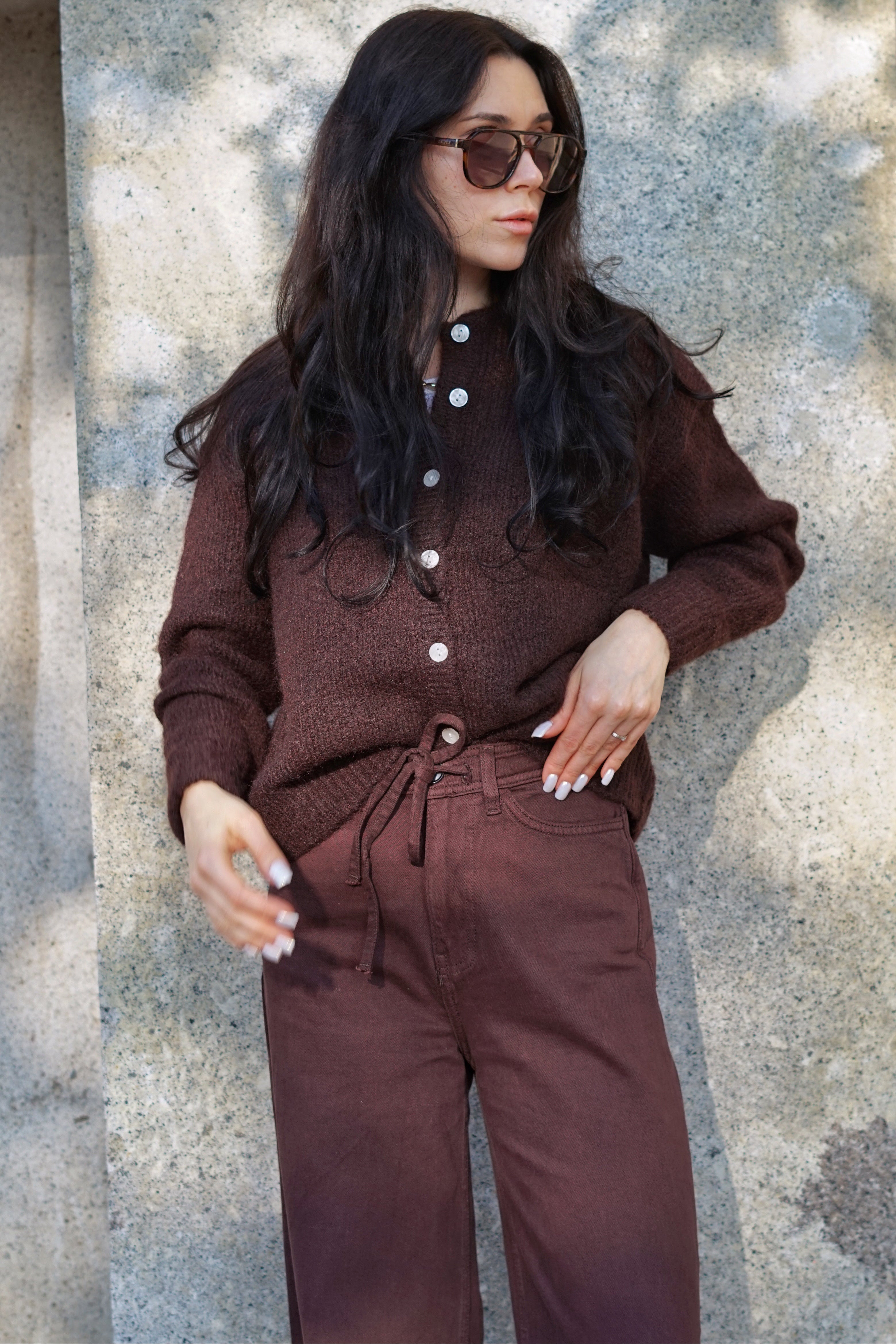 A woman wearing a matching brown knit cardigan and drawstring pants set, styled with dark sunglasses, posing against a textured concrete wall with natural dappled shadows.