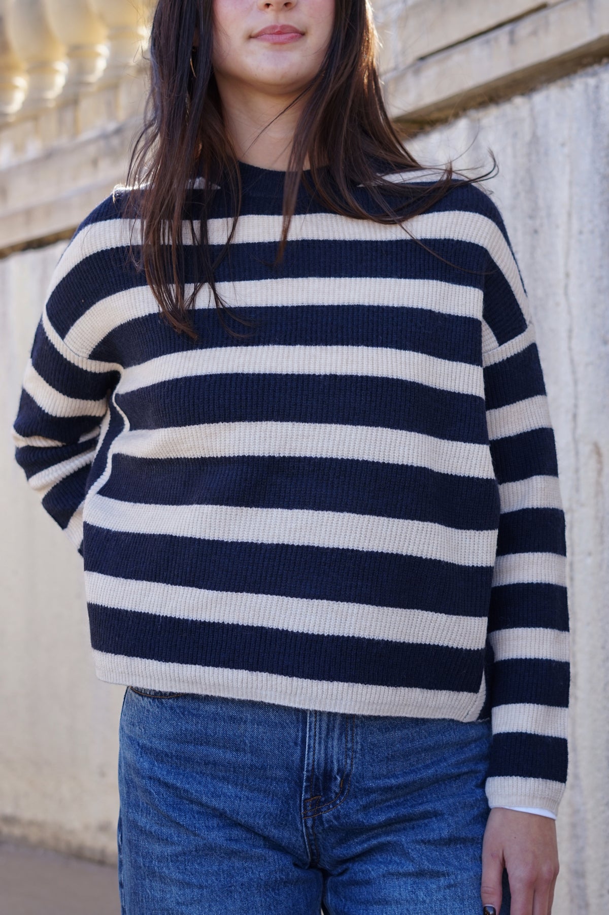 A woman wearing a navy and cream horizontally striped knit sweater with blue jeans, standing against a light-colored stone wall.