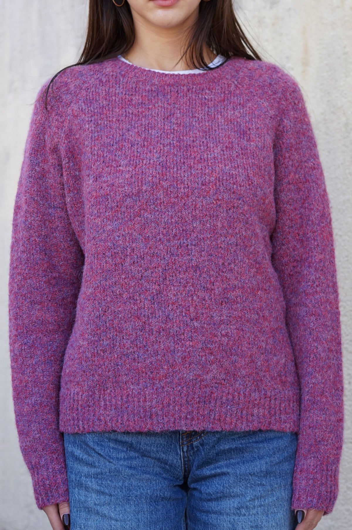 A woman wearing a chunky knit crewneck sweater in a heathered purple and pink colorway, paired with blue jeans, standing against a neutral textured wall.