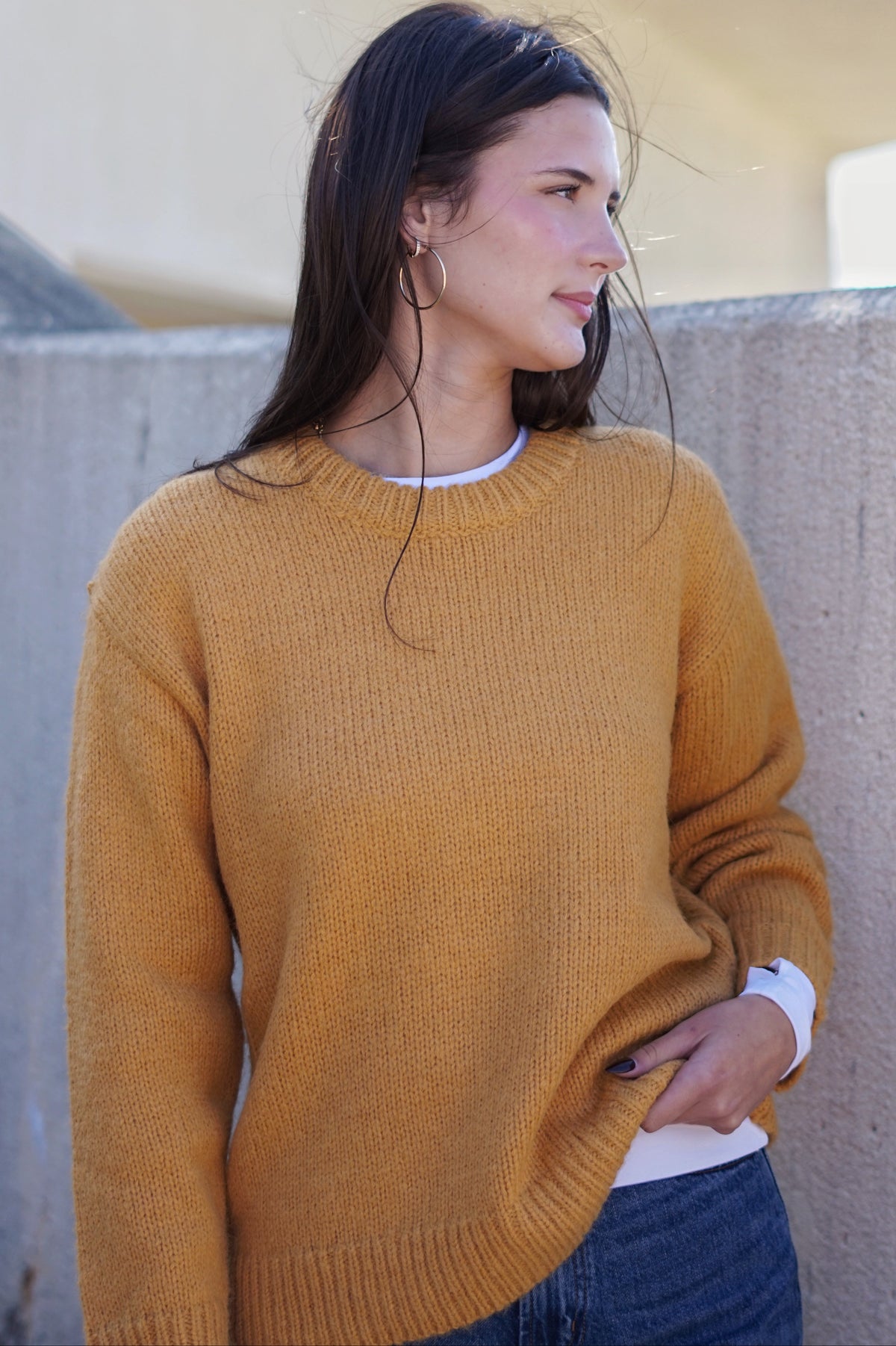 A woman wearing a mustard yellow knit crewneck sweater over a white undershirt and blue jeans