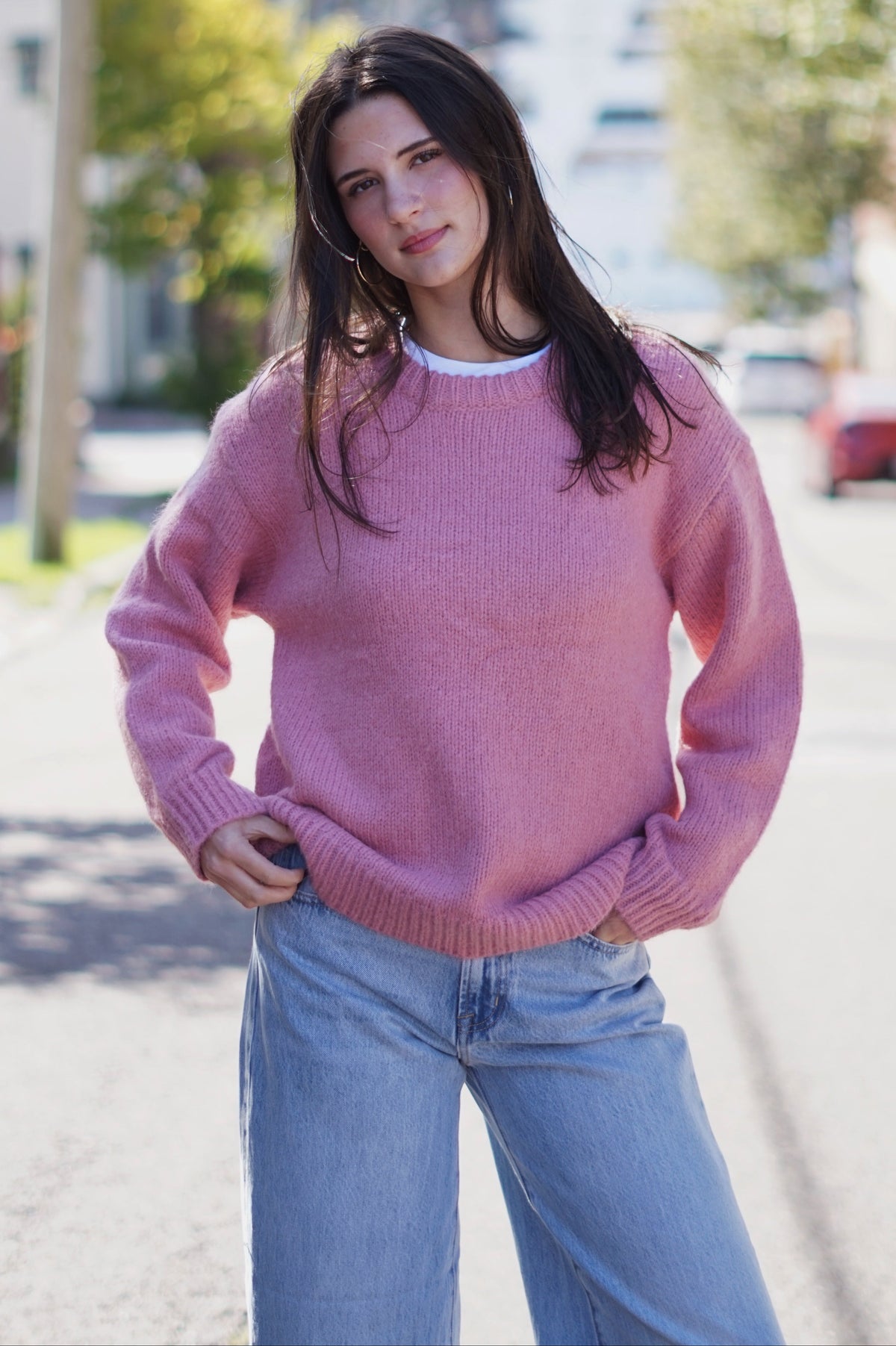 A woman wearing a pink knit crewneck sweater and light blue jeans standing on a sunny residential street.