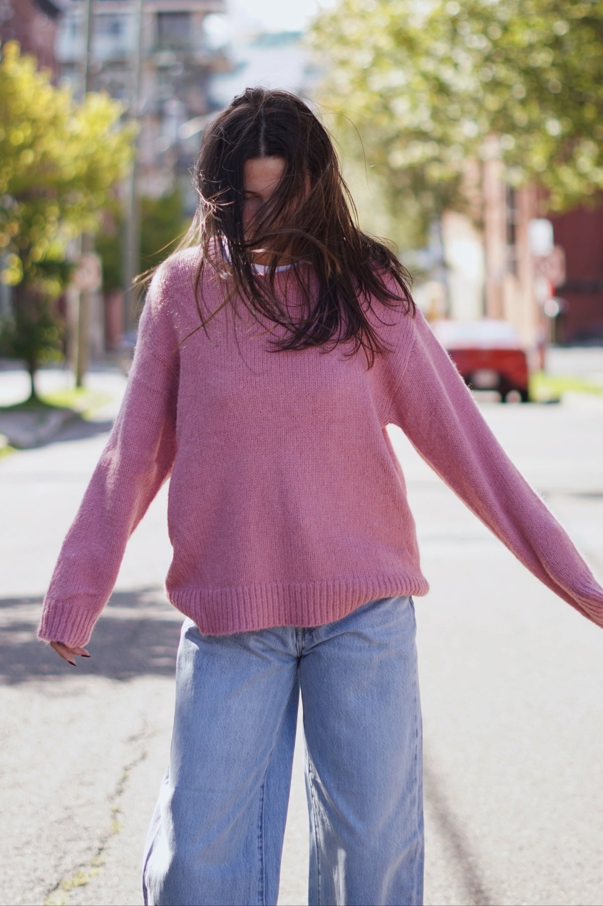 A woman wearing a dusty pink knit sweater and light blue wide-leg jeans walking down a sunlit residential street.