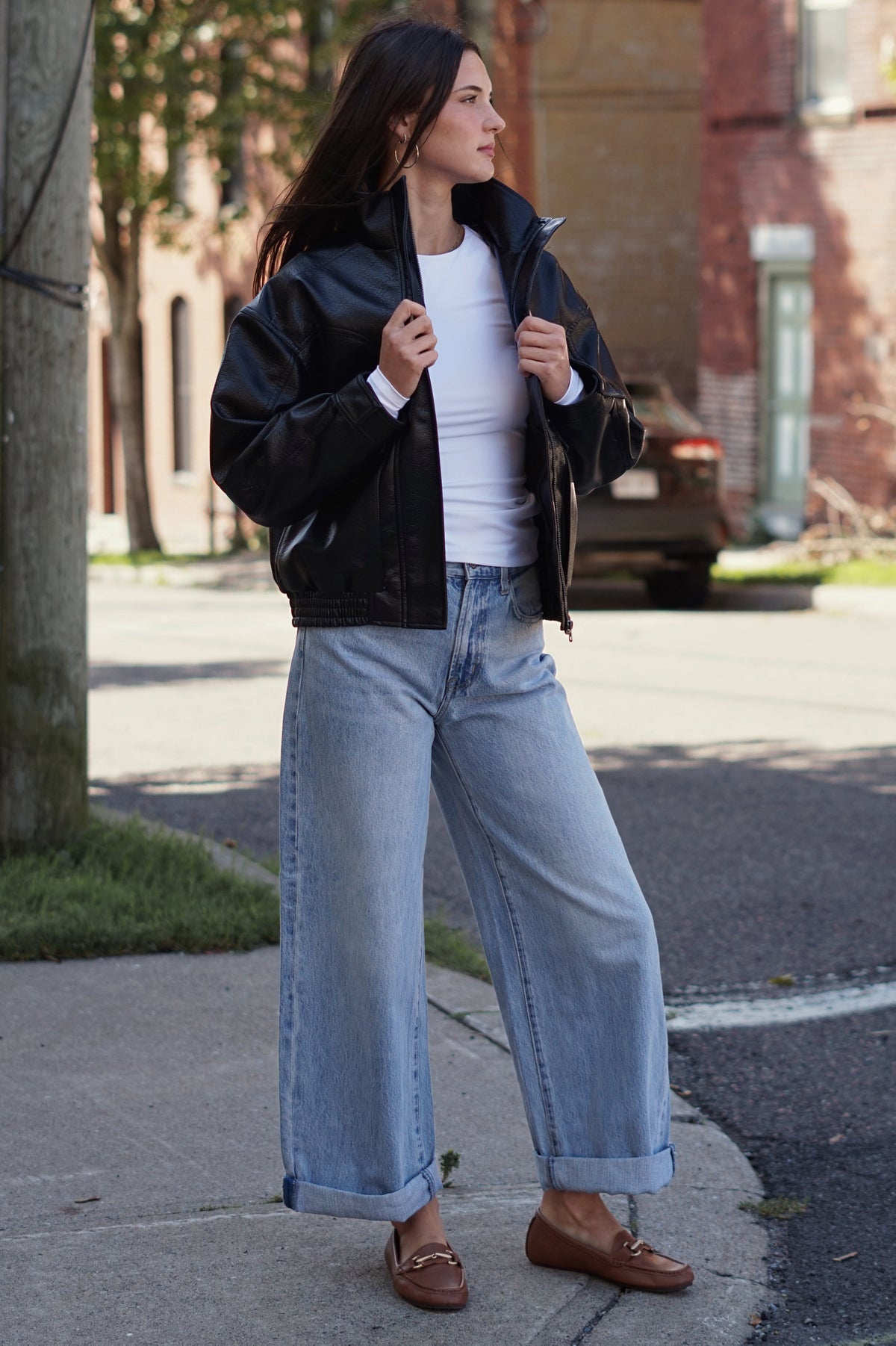 A woman wearing a black leather jacket, white top, wide-leg light blue jeans, and brown loafers