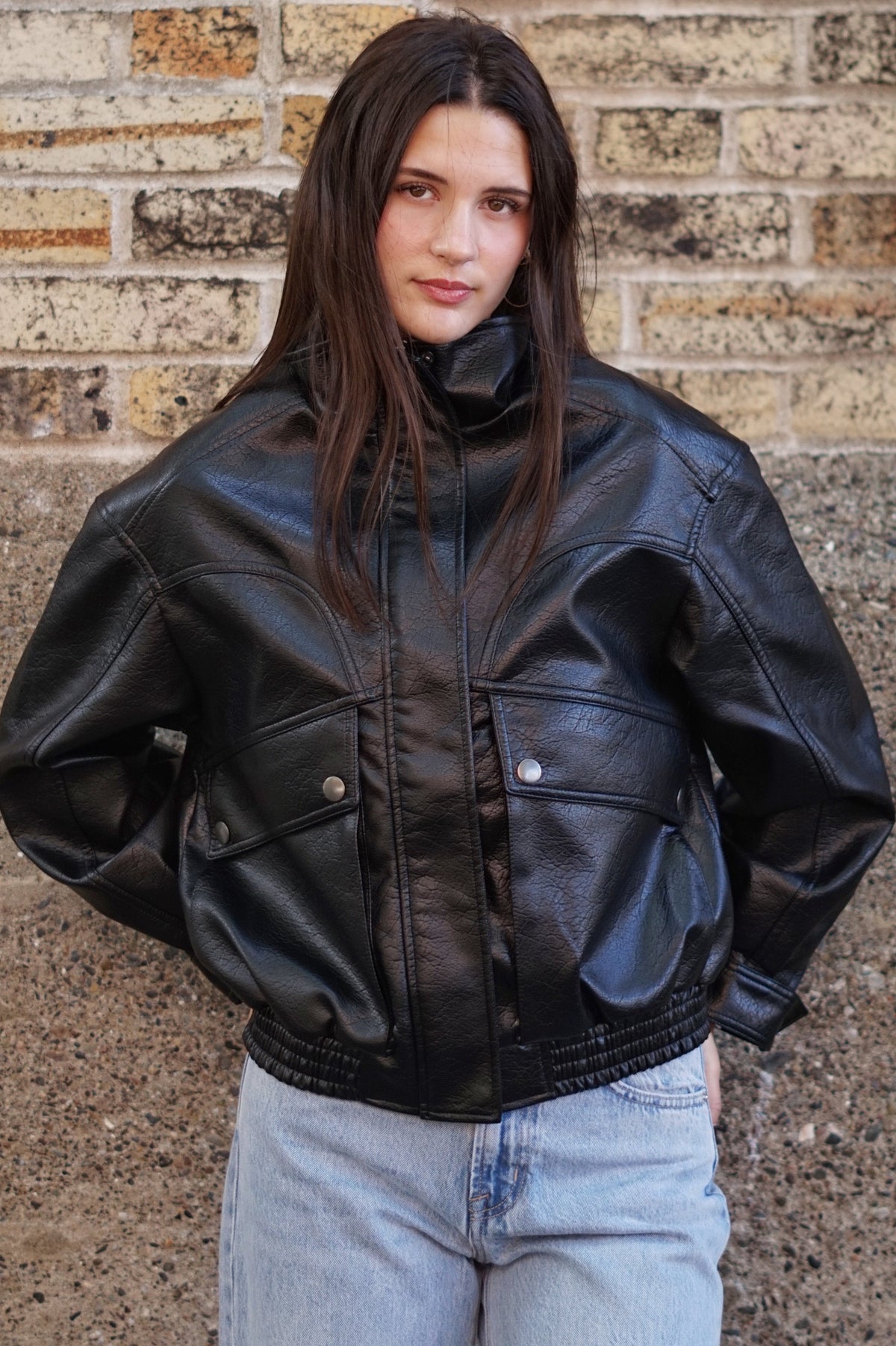 A woman with long dark hair stands in front of a brick wall wearing a black leather jacket and light-wash blue jeans.