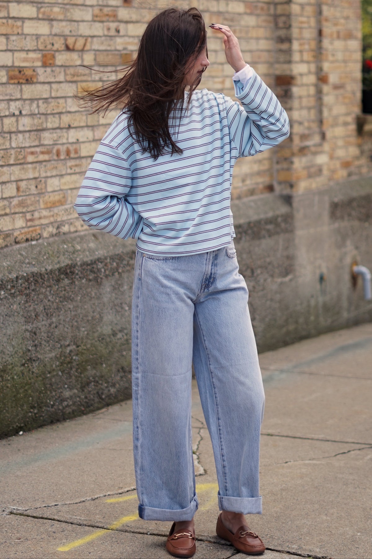 A woman wearing a light blue and white striped long-sleeve top, light-wash wide-leg jeans, and brown leather loafers.