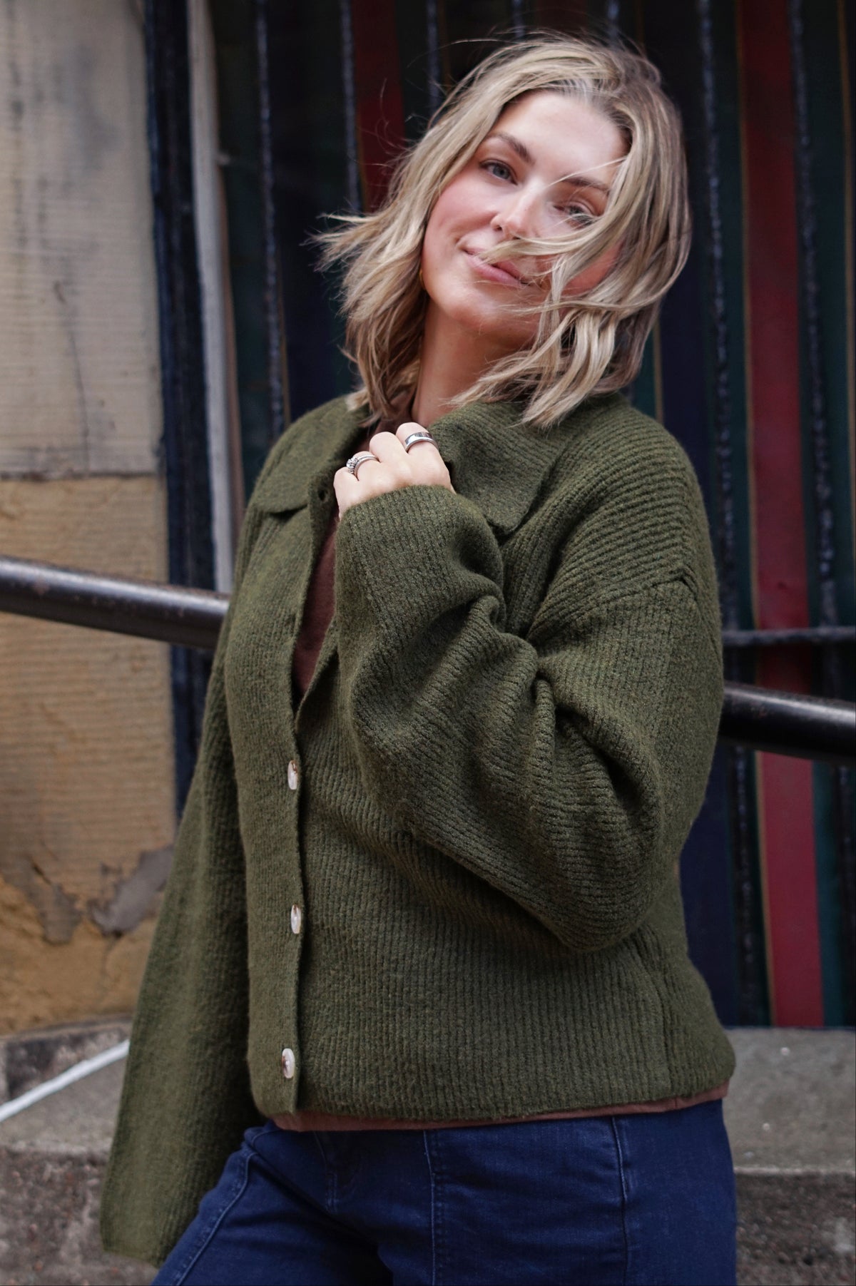 A woman wearing a ribbed olive green button-up cardigan over a brown top and blue jeans, standing outdoors in front of a dark metal gate.