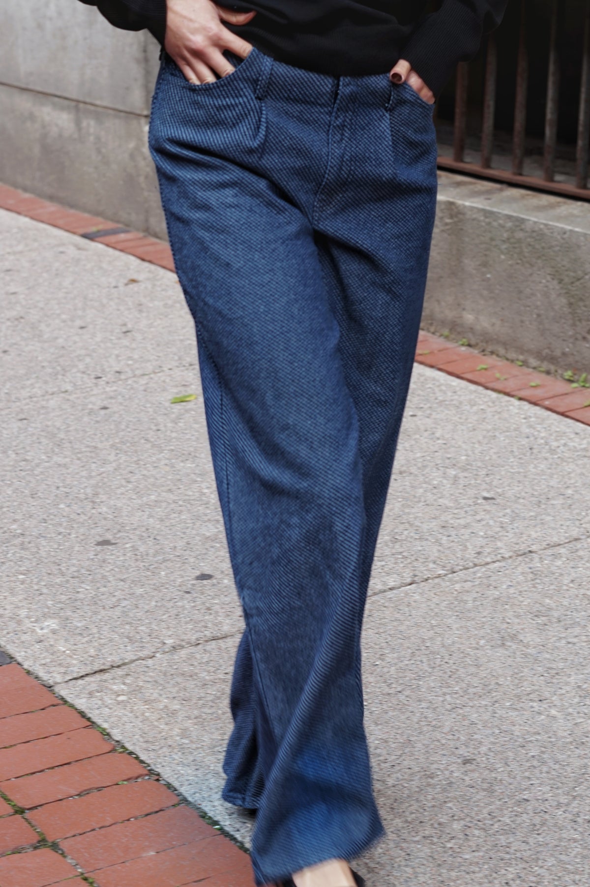 A person wearing wide-leg blue jeans with a subtle herringbone pattern, standing on a concrete sidewalk with brick borders.