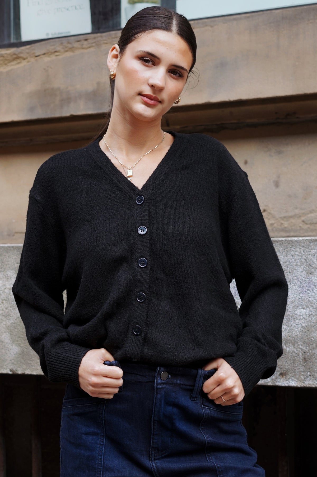 A woman wearing a black V-neck button-up cardigan sweater and dark blue jeans, standing outdoors in front of a stone building.