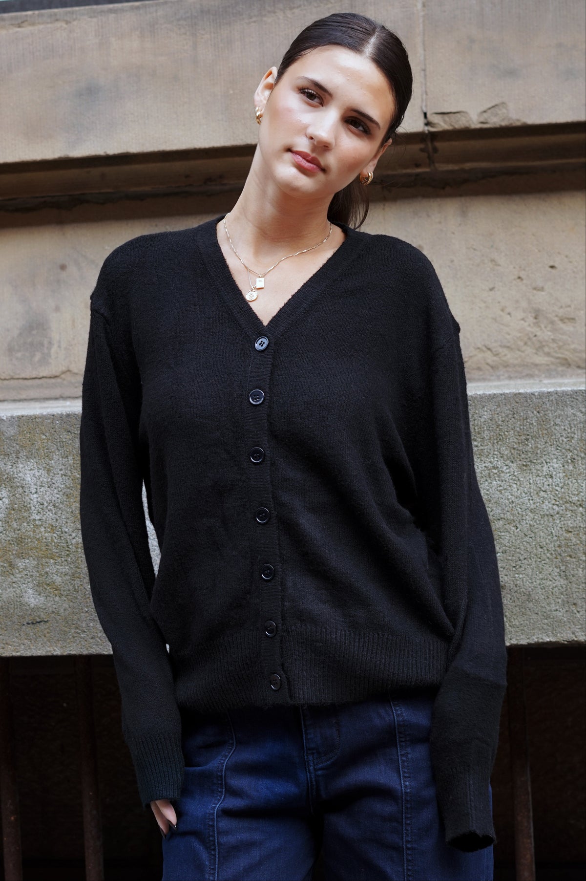 A woman wearing a black button-up cardigan sweater and dark blue jeans, standing outdoors against a stone wall.