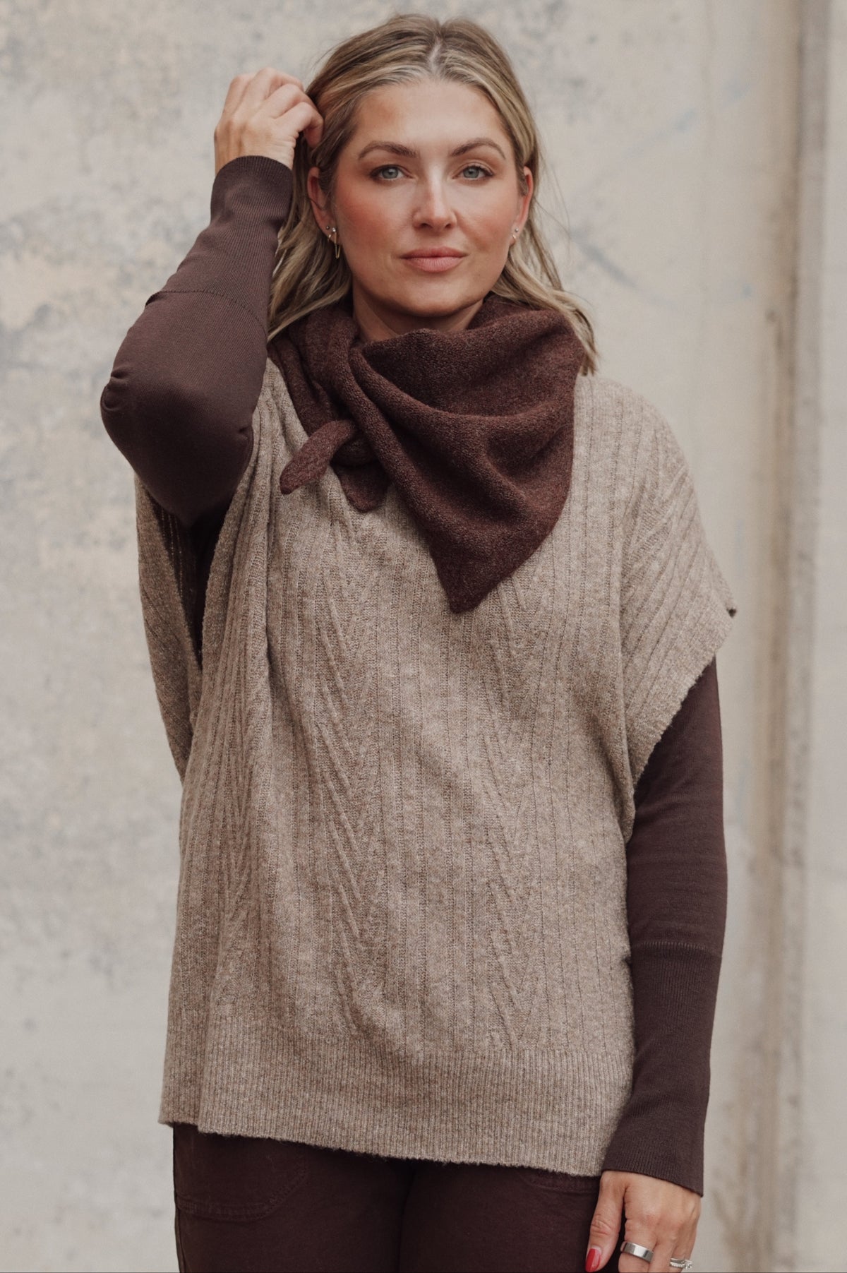 A woman wearing a taupe cable-knit sleeveless sweater vest over a dark brown long-sleeve top and matching brown pants, styled with a dark brown knit cowl scarf, standing against a textured concrete wall.