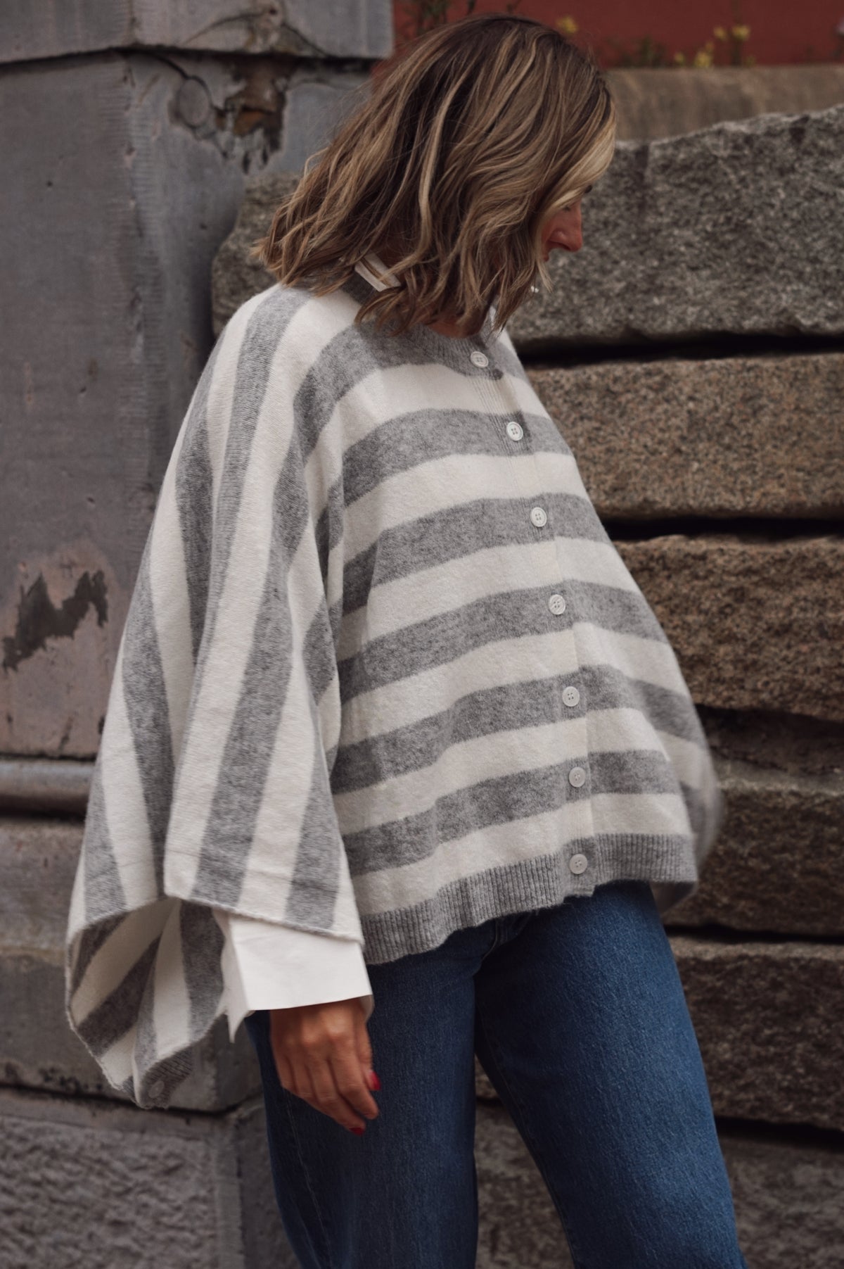 A woman wearing a gray and off-white striped knit poncho sweater with a button-down front, paired with blue jeans, standing against a rustic stone wall.