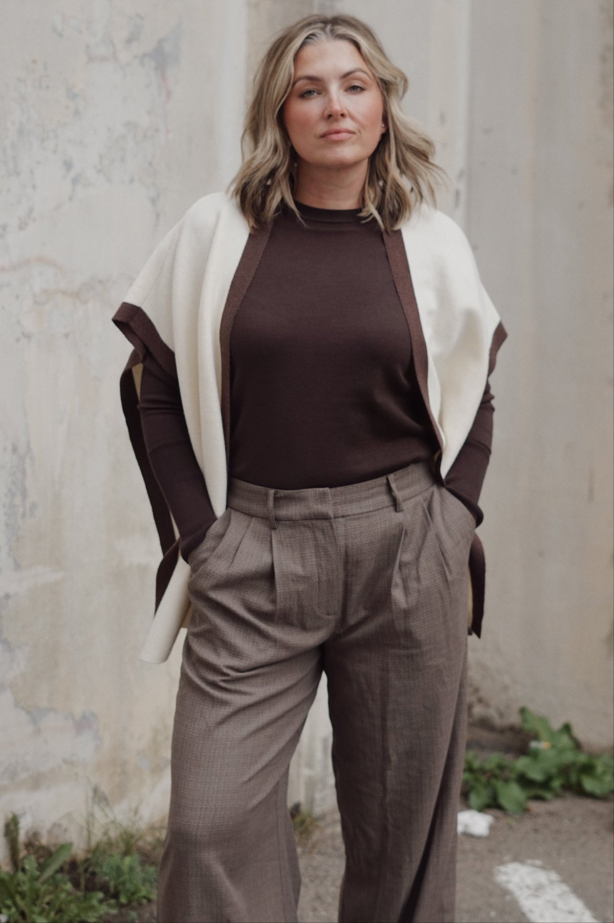 A woman stands outdoors against a textured concrete wall wearing a dark brown ribbed knit top, high-waisted taupe wide-leg trousers, a cream draped cardigan, and a dark brown belt.