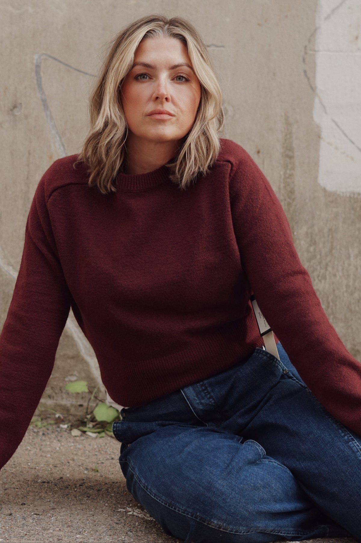 A woman with blonde hair sits on a concrete surface wearing a dark burgundy knit sweater and blue jeans against a textured concrete wall.