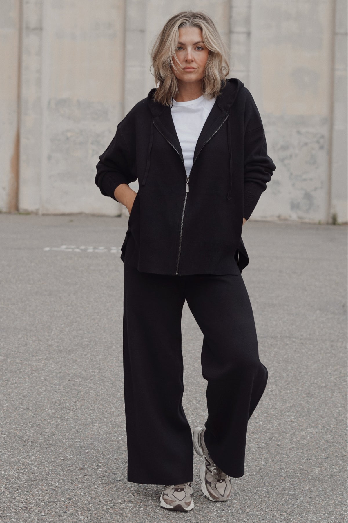 A woman stands outdoors wearing a black zip-up hoodie, black wide-leg sweatpants, a white t-shirt, and gray sneakers against a concrete wall background.