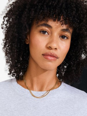 A close-up portrait of a woman with curly black hair wearing a light gray crewneck shirt and a delicate gold chain necklace, set against a dark background.