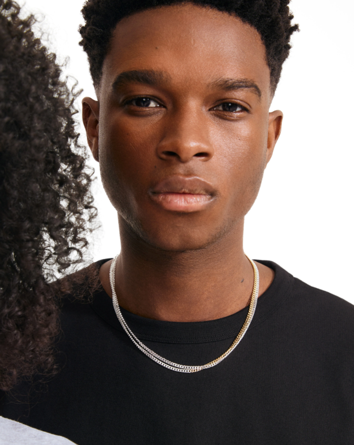 A close-up portrait of a man wearing a delicate silver chain necklace over a black t-shirt, with a white background and another person partially visible on the left.