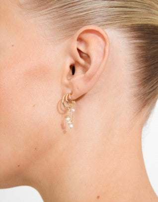 A close-up of a model's ear wearing a delicate gold hoop earring with dangling pearl-like beads.