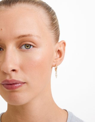 A close-up of a woman's face and ear wearing a delicate gold dangle earring with small clear stones, against a plain white background.