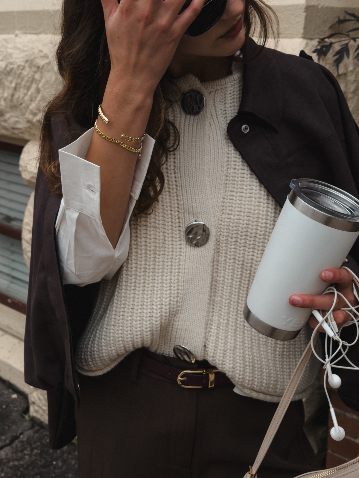 A person wearing a cream ribbed knit sweater vest over a white button-down shirt, with a dark brown jacket and brown trousers, holding a white insulated travel tumbler with earphones in a street setting.