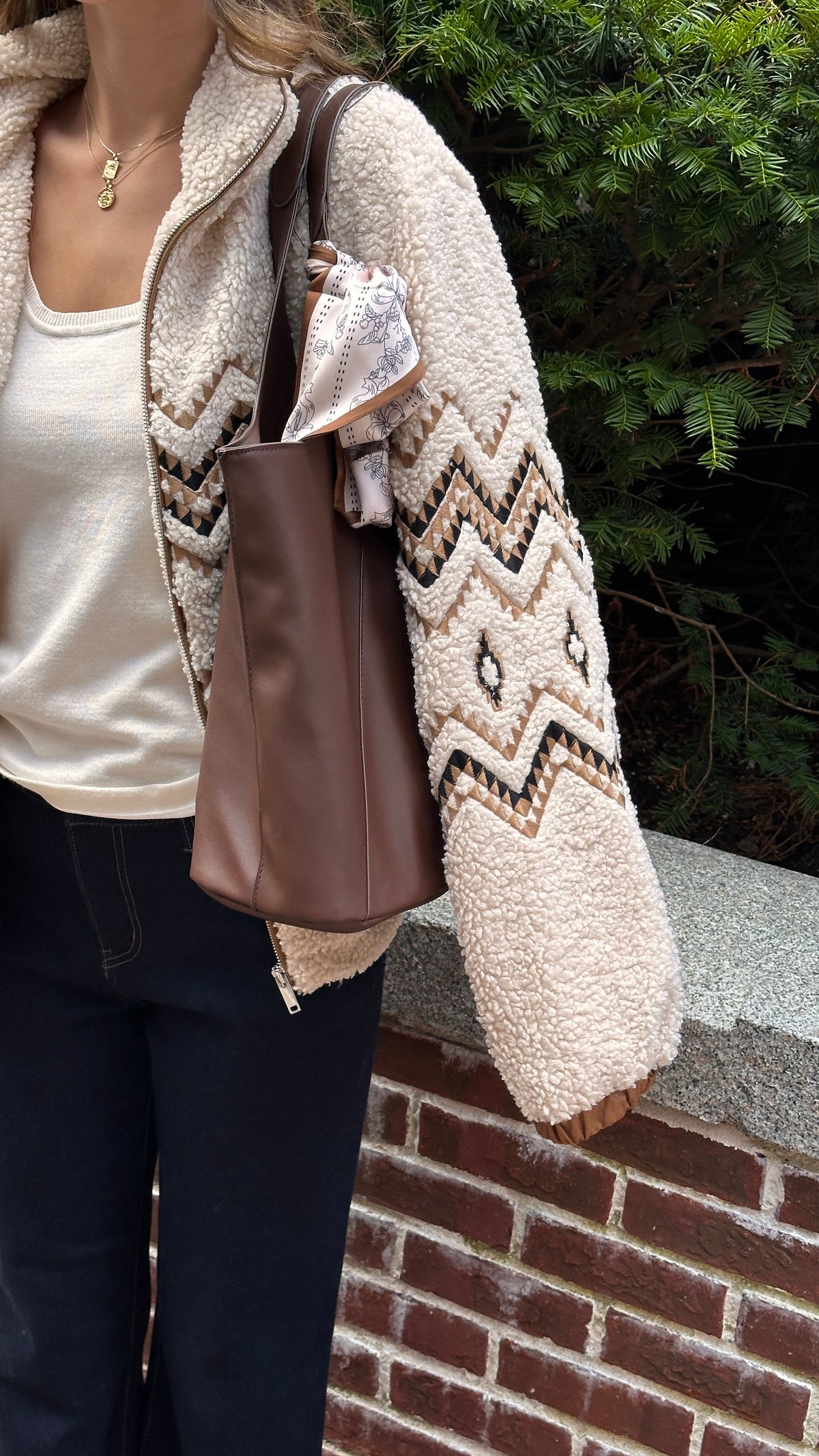 Close-up of a cream sherpa jacket with brown geometric detailing styled with a white top, dark jeans and a brown shoulder bag with a printed scarf tied to the handle.