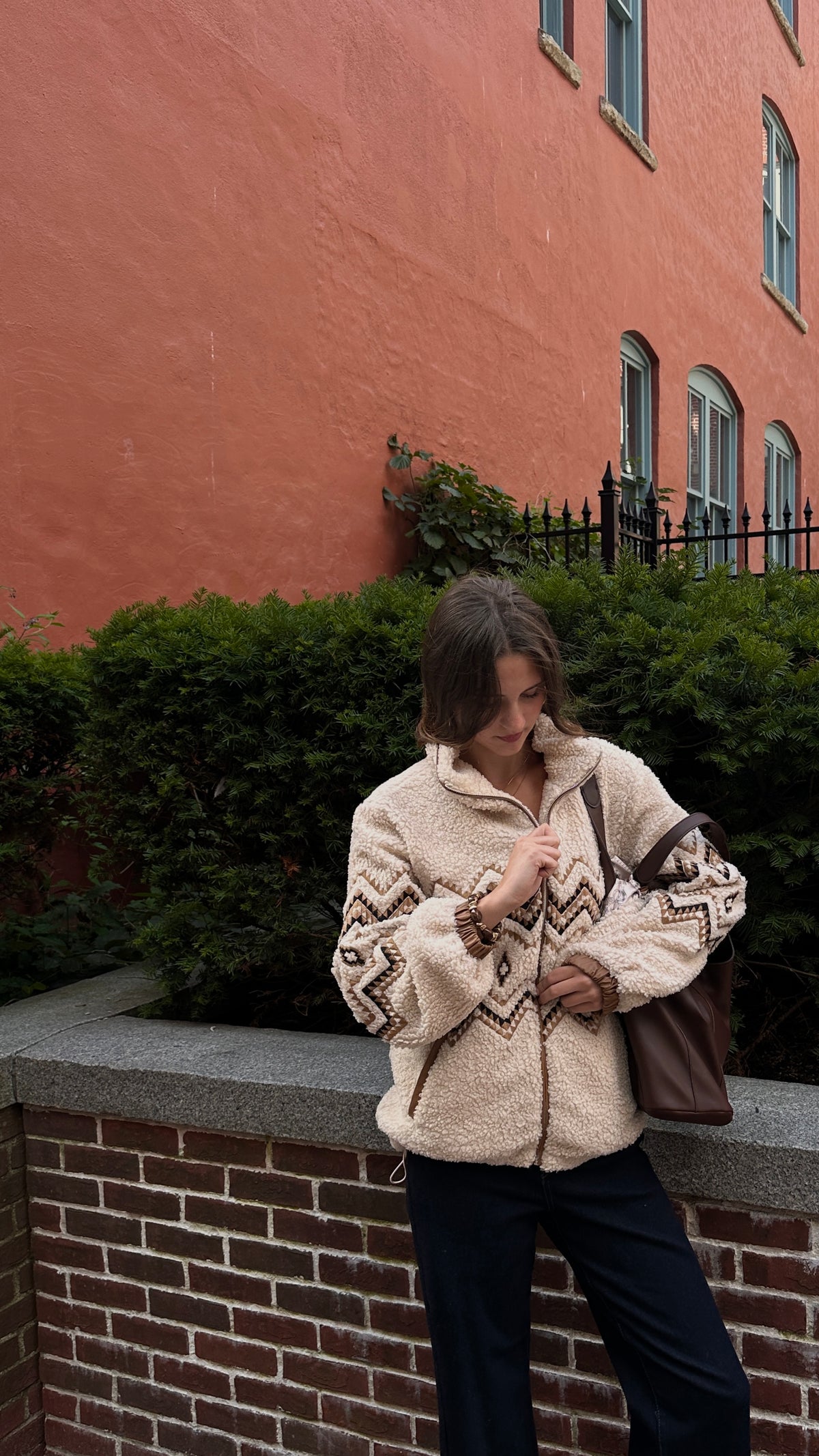 Woman wearing a cream sherpa jacket with brown geometric detailing, dark jeans and a brown shoulder bag, posing outdoors in front of greenery and a red brick building.