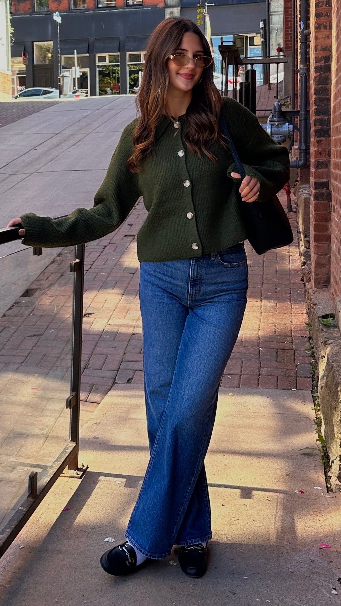 Woman wearing a dark green button-front cardigan with blue jeans, sunglasses and a black shoulder bag on a brick-lined city street.