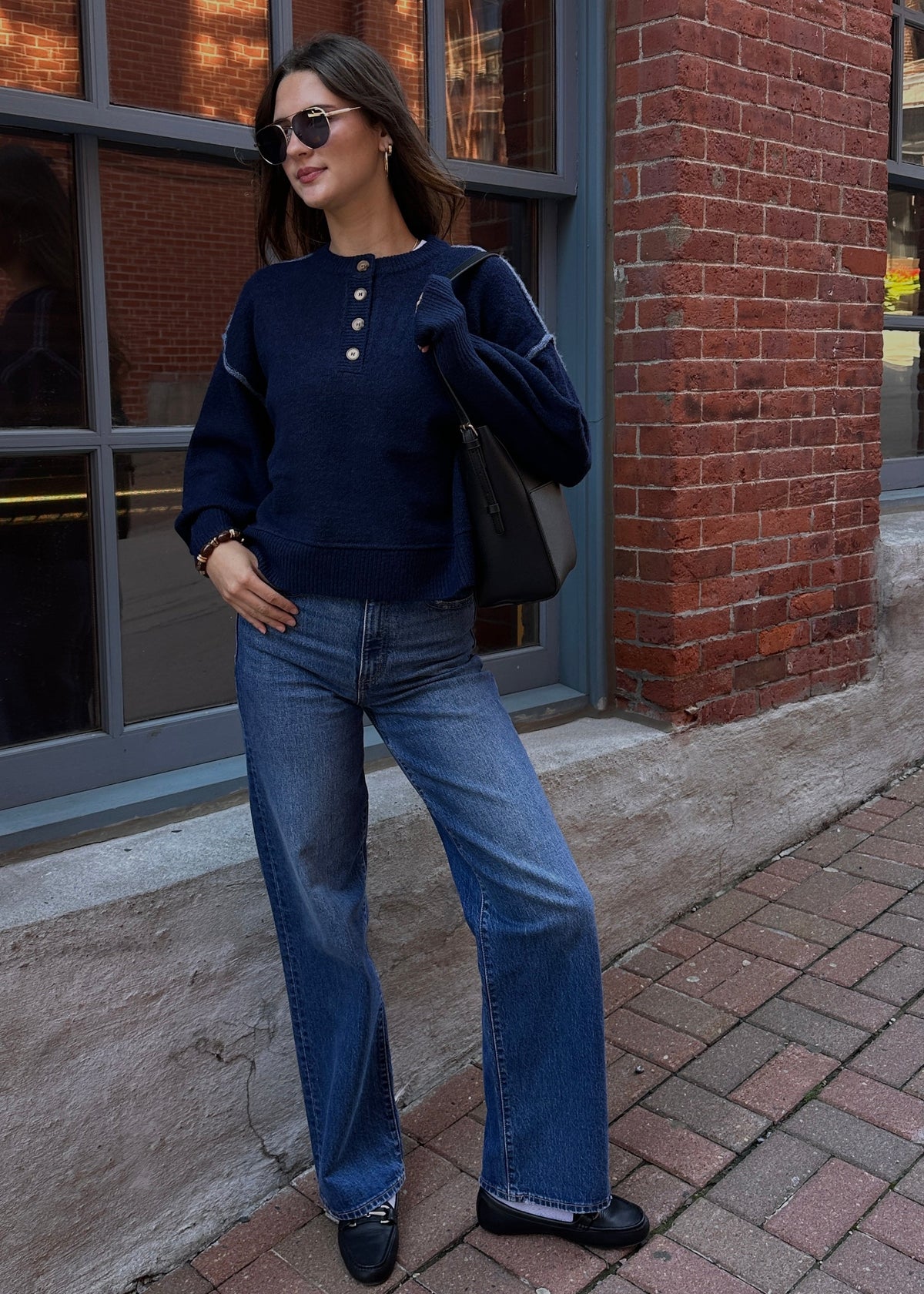 Woman wearing a navy button-detail sweater with wide-leg blue jeans, black shoes and sunglasses, standing outside against a red brick building.