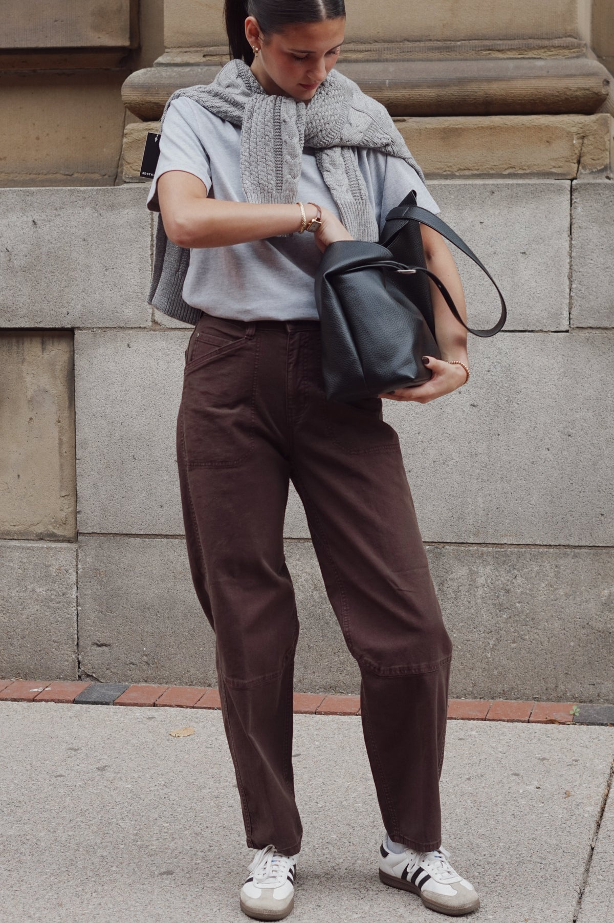 A woman wearing a light gray t-shirt, a chunky gray knit scarf, dark brown wide-leg pants, and white sneakers with black stripes, holding a black leather shoulder bag against a stone wall background.